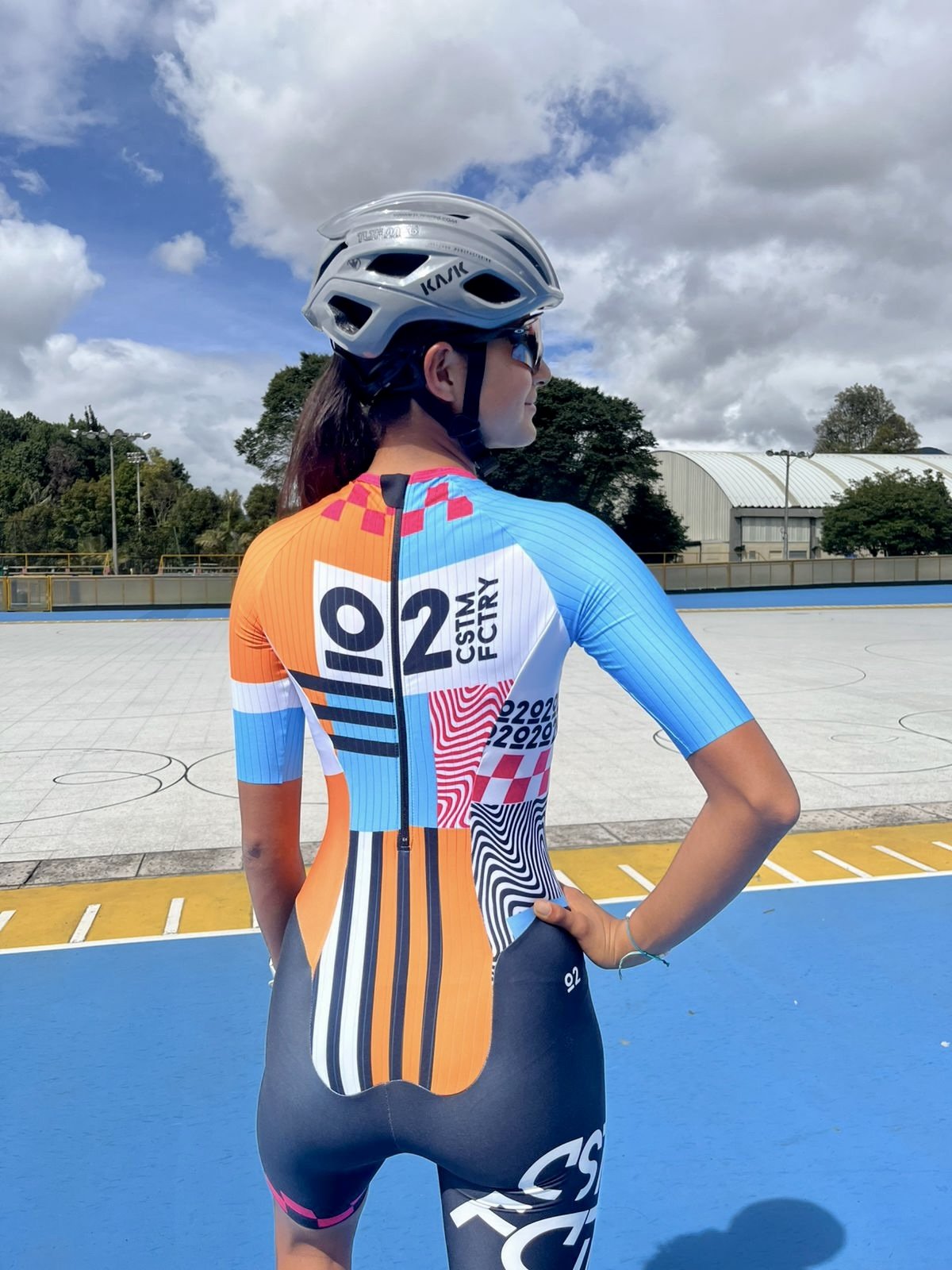 A young female roller skater wearing a blue, yellow, and red Colombia sports uniform, a white helmet, and reflective sunglasses is skating on a blue and orange outdoor rink with trees and a cloudy sky in the background.