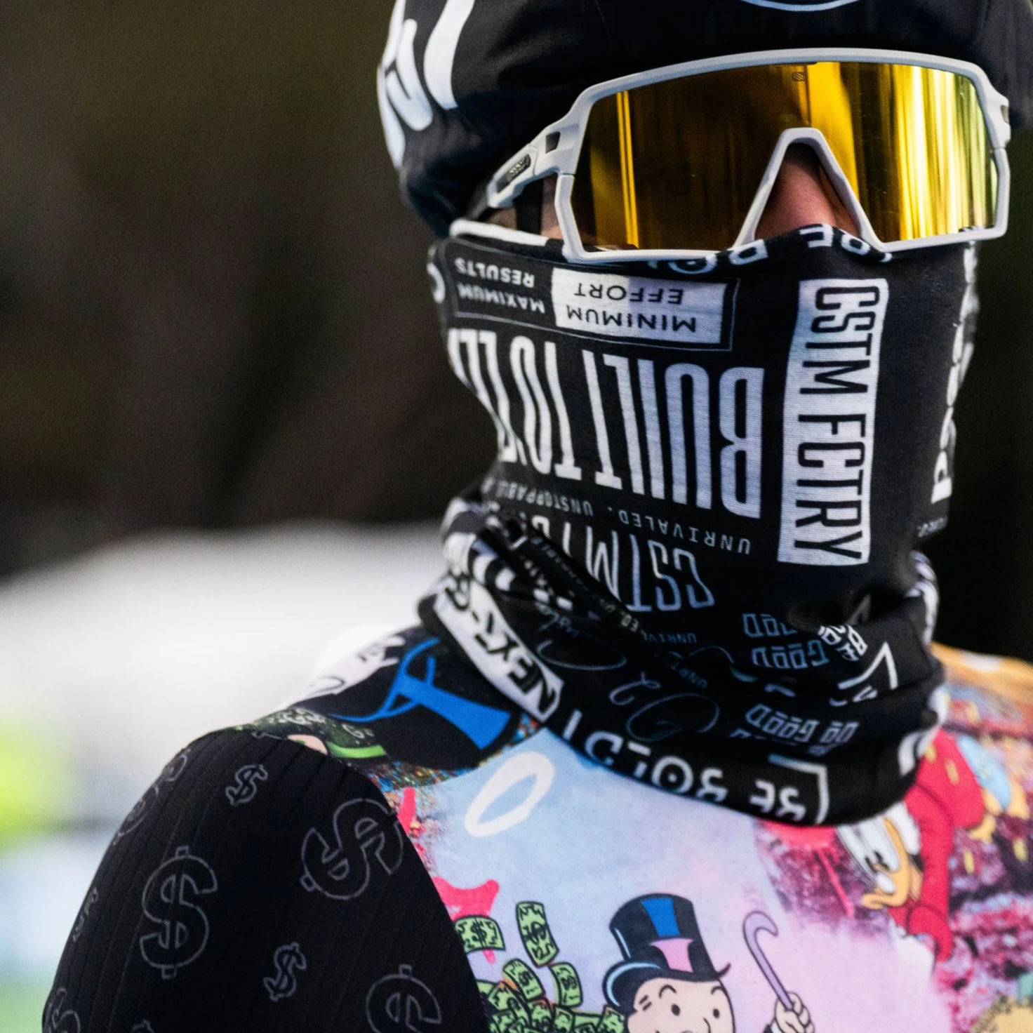 A young man wearing a black helmet, red sunglasses, a black sleeveless athletic top, and black shorts with dollar signs, sitting on a bench outdoors in a park with green grass and a yellow fence.