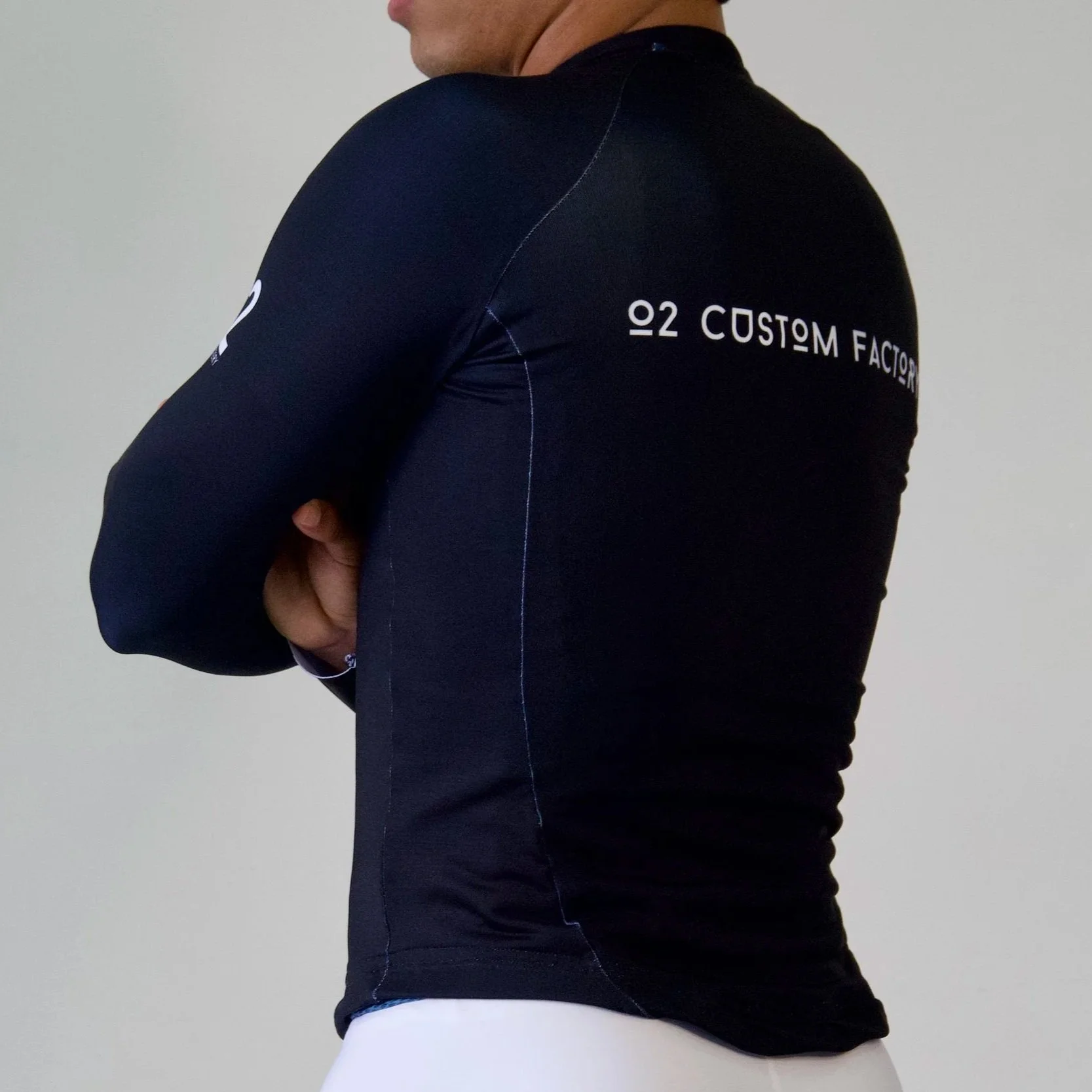 A person wearing a black athletic zip-up jacket with white 'O2' and 'Custom Factory' logos on the chest and sleeve, standing with arms crossed against a plain wall.