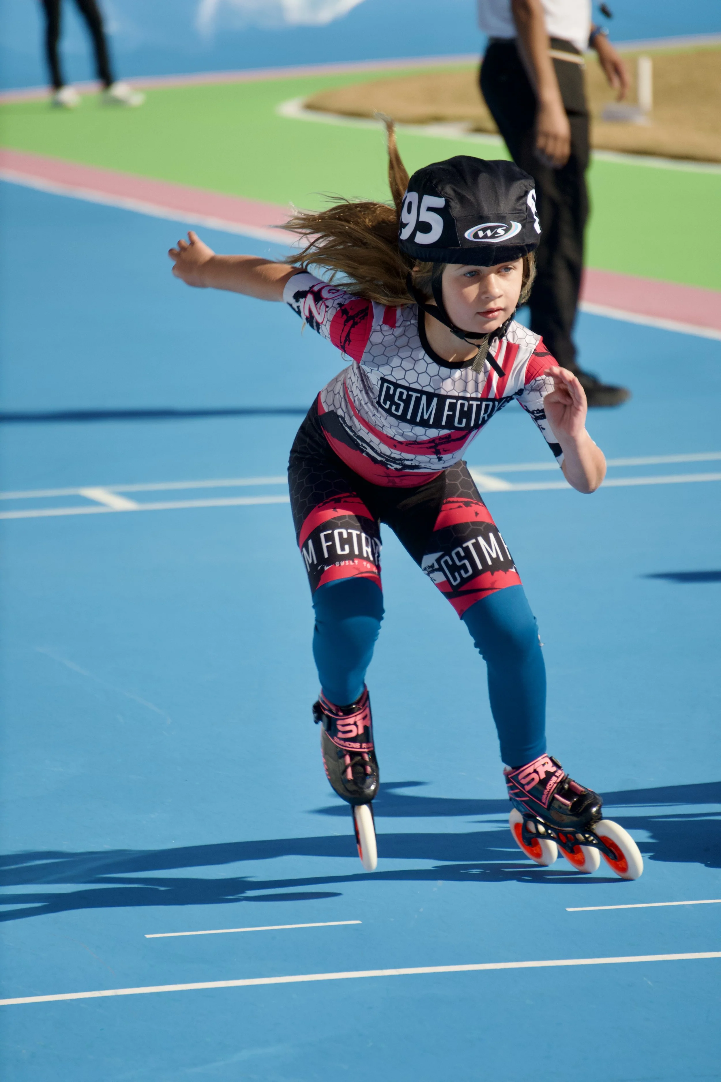 A young female roller skater wearing a blue, yellow, and red Colombia sports uniform, a white helmet, and reflective sunglasses is skating on a blue and orange outdoor rink with trees and a cloudy sky in the background.