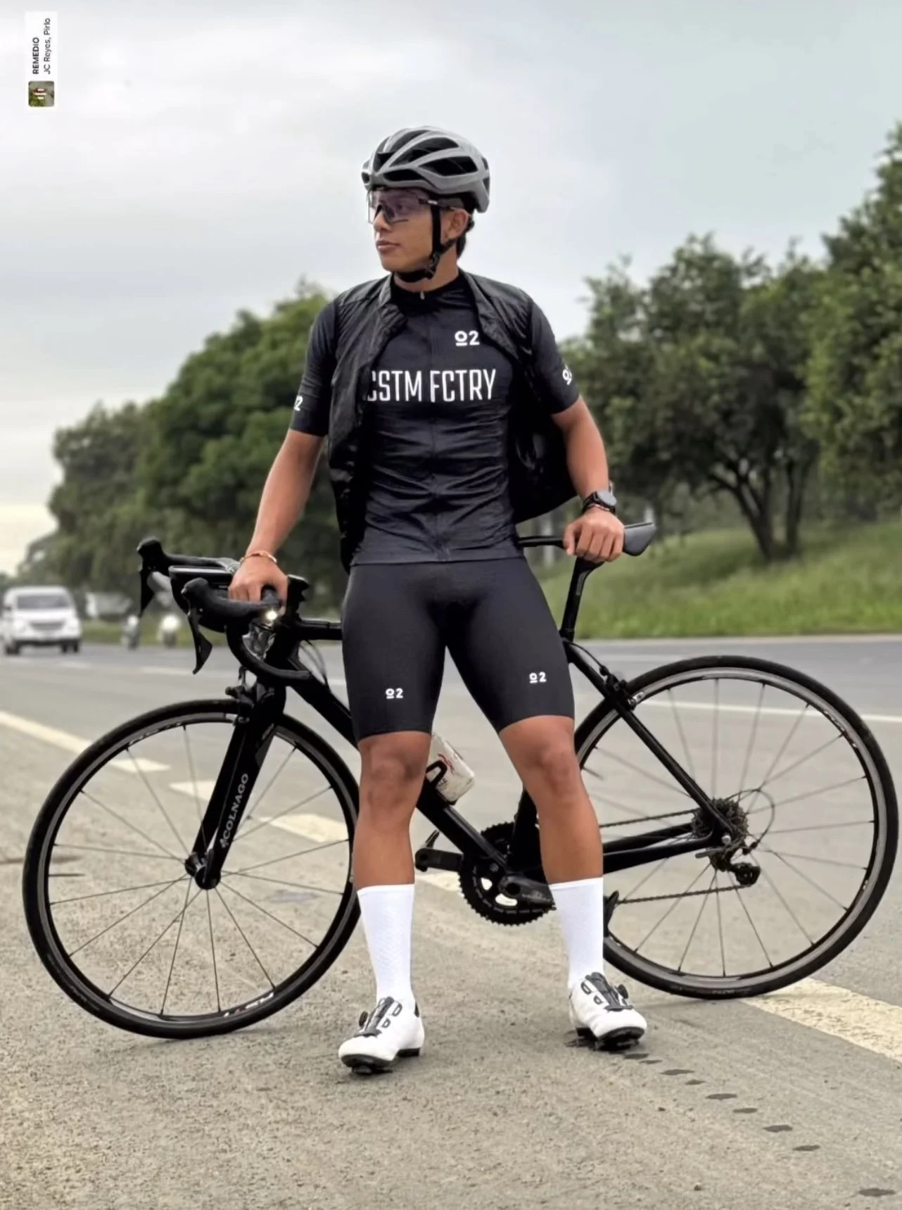 A woman stands with a black road bike on the side of a highway, wearing a black helmet, sunglasses, black cycling suit, white socks, and cycling shoes, with trees and cars in the background.