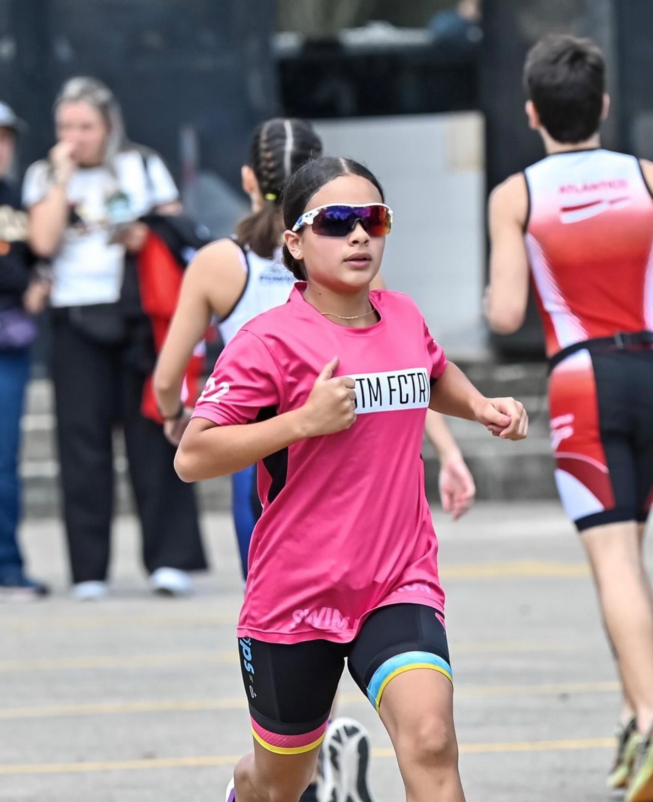 A woman wearing a black cycling suit and a white and black helmet standing outdoors near a skate park or similar area, looking away with her hand on the back of her head, with trees and buildings in the background.