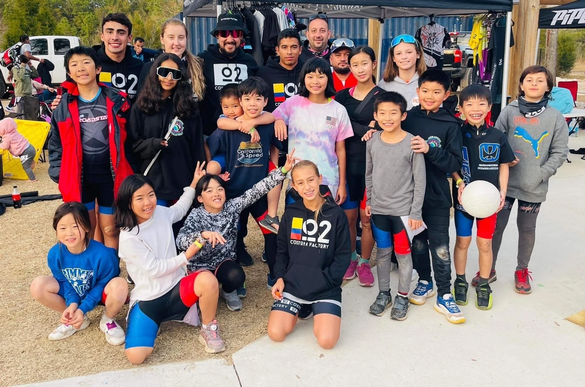 Group of young kids and adults gathered outdoors at a sports or athletic event, with some wearing sports gear, holding a volleyball, and smiling at the camera.