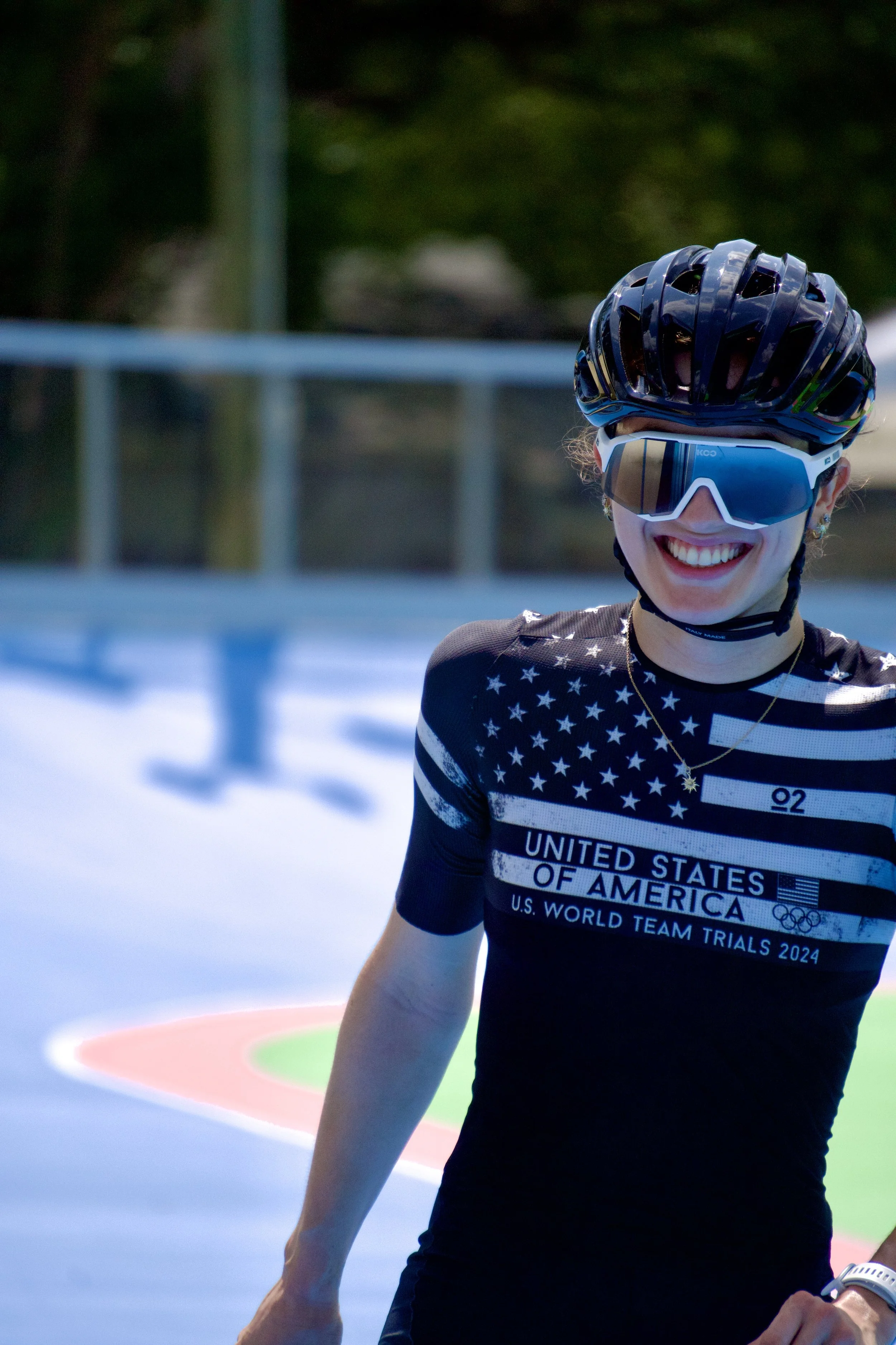 Smiling female athlete wearing a helmet, sunglasses, and a black sports jersey with American flag and United States of America text, standing on a sports track or field.