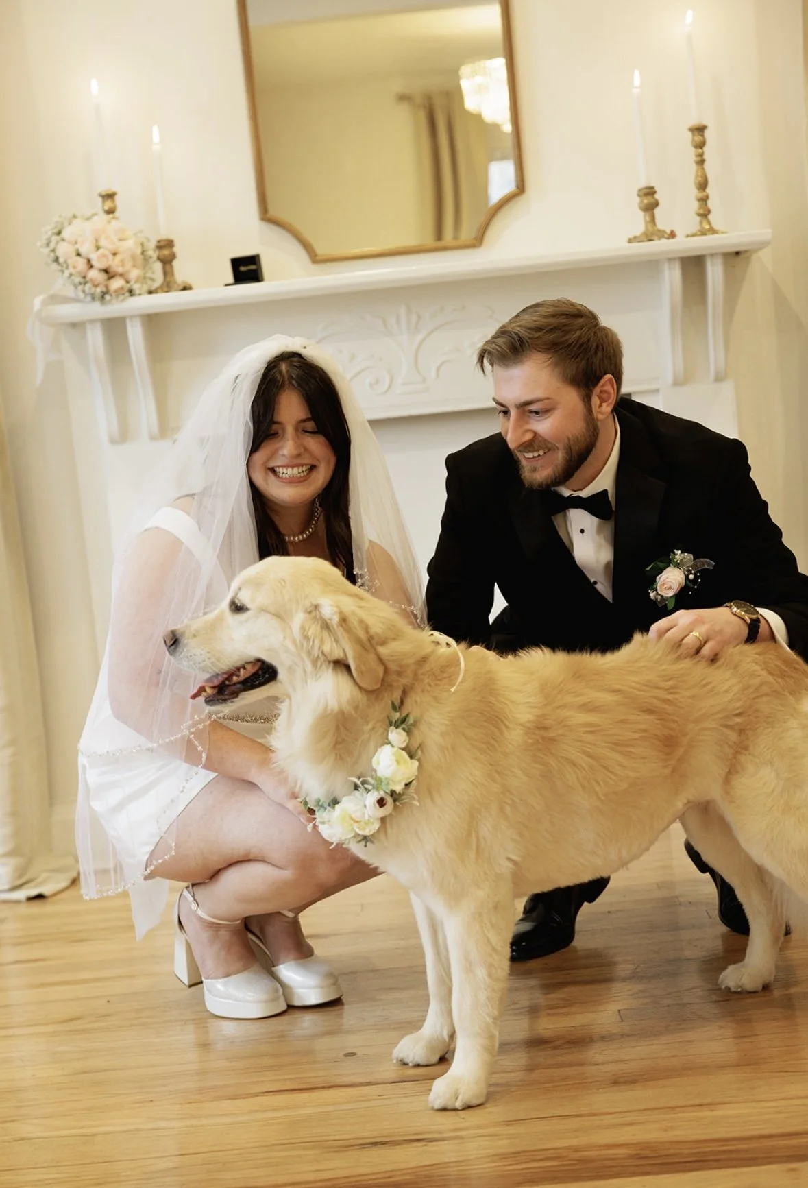 A bride and groom smiling happily with a golden retriever dog during a wedding celebration in a decorated indoor setting.