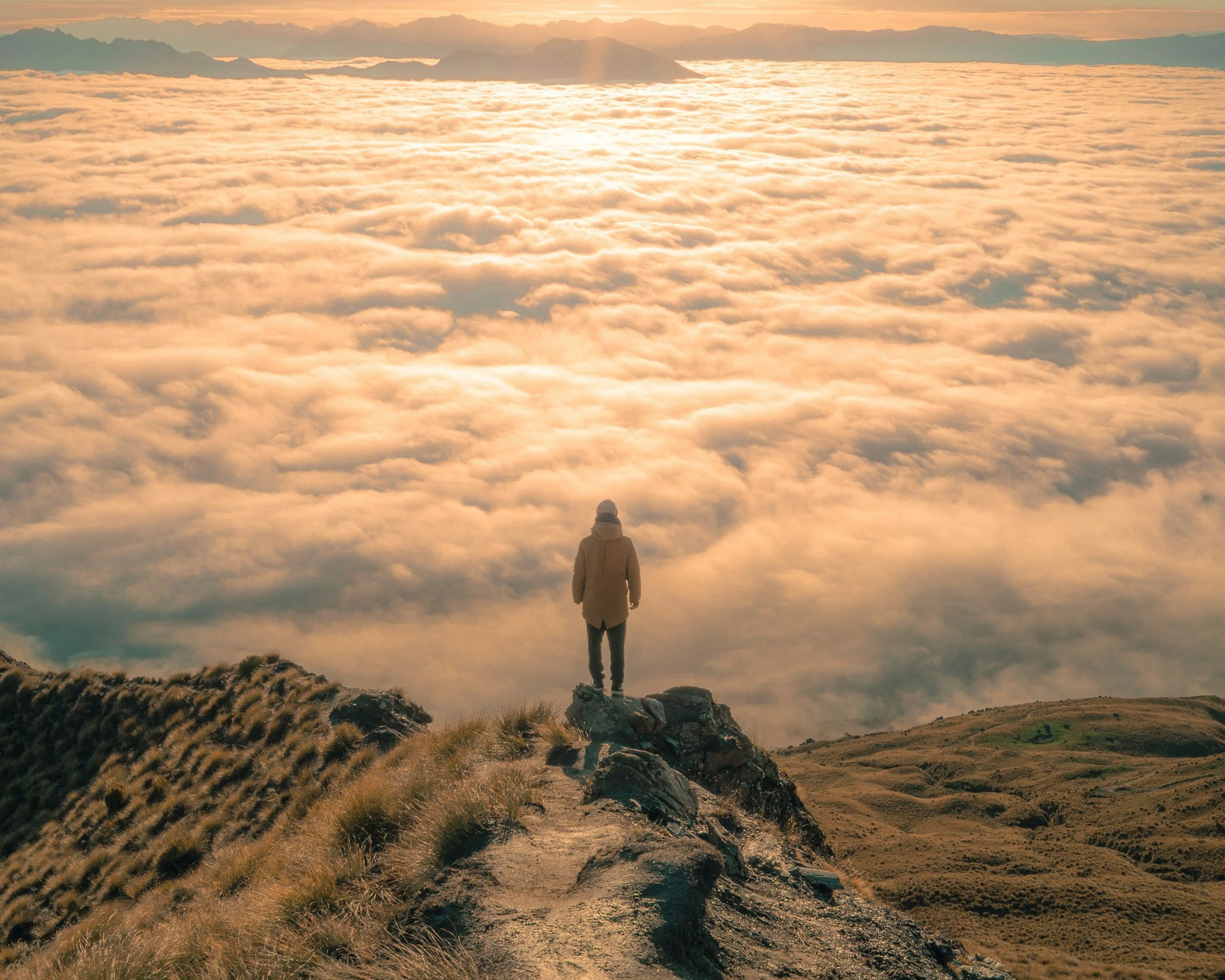 Person standing on a mountain trail overlooking clouds and distant mountains at sunset.