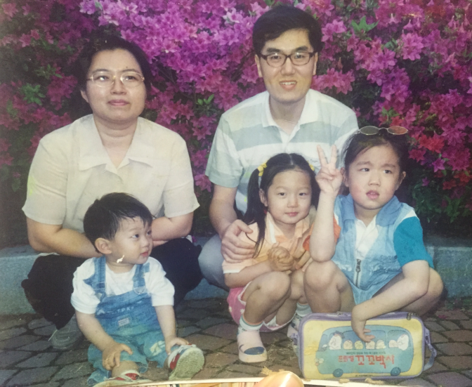 Family with three children posing outdoors in front of blooming flowers, two adults sitting behind, and the children in front, one holding a box.