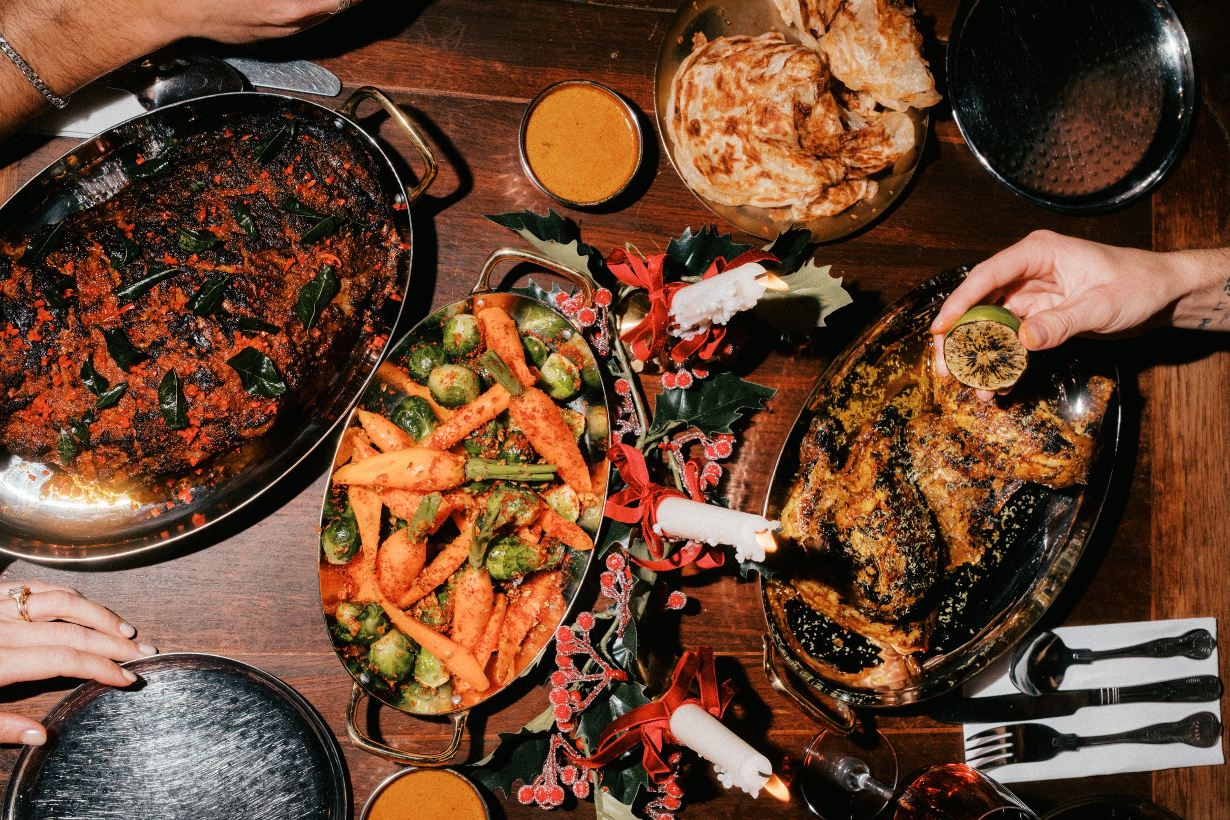 A festive holiday meal on a wooden table with various dishes including roasted meat, vegetables, sauces, and bread, decorated with Christmas-themed decor.
