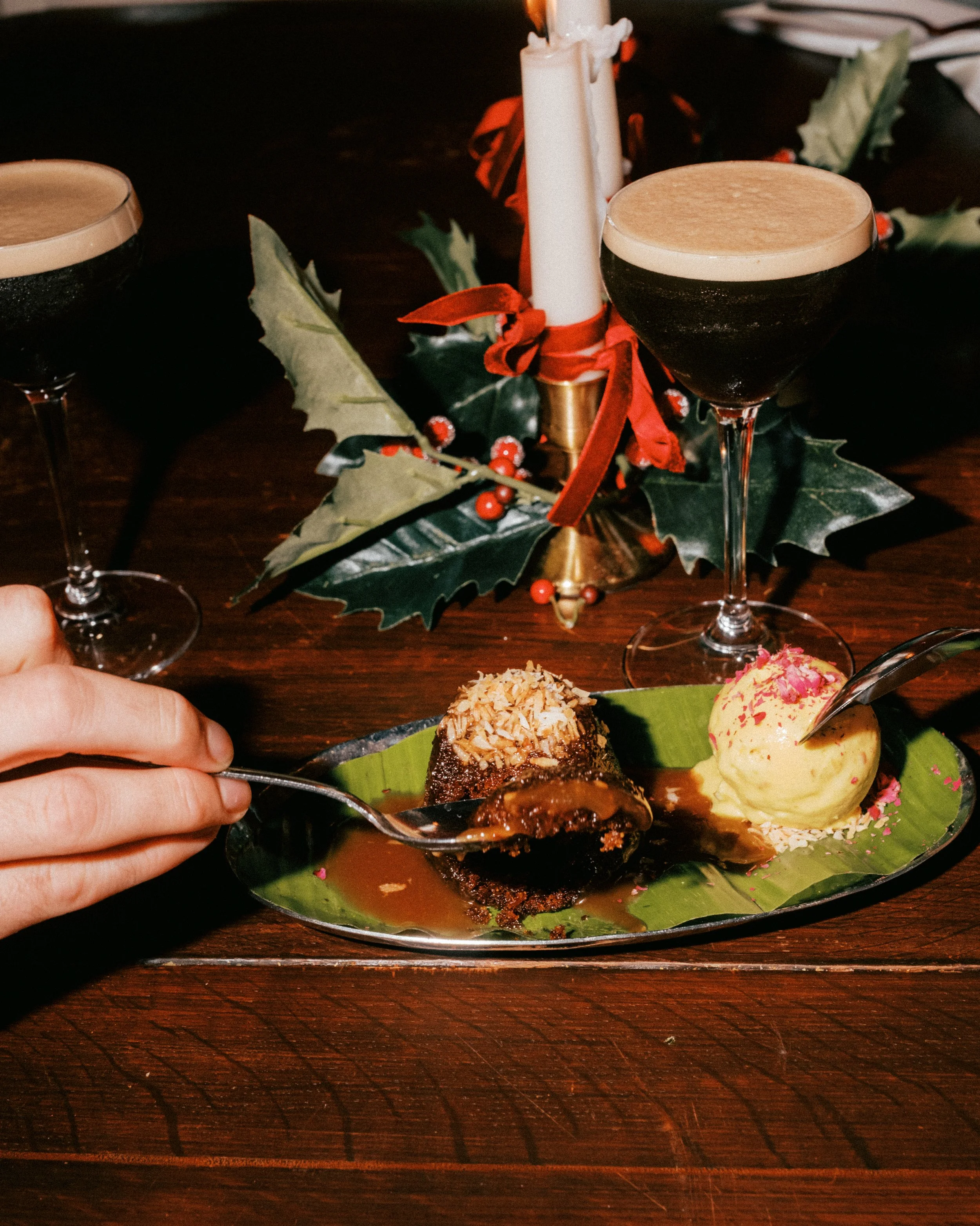 A holiday dessert plate with ice cream, decorated cake, and a chocolate cake slice, served on a green leaf, with holiday decorations and two glasses of dark beer in the background.