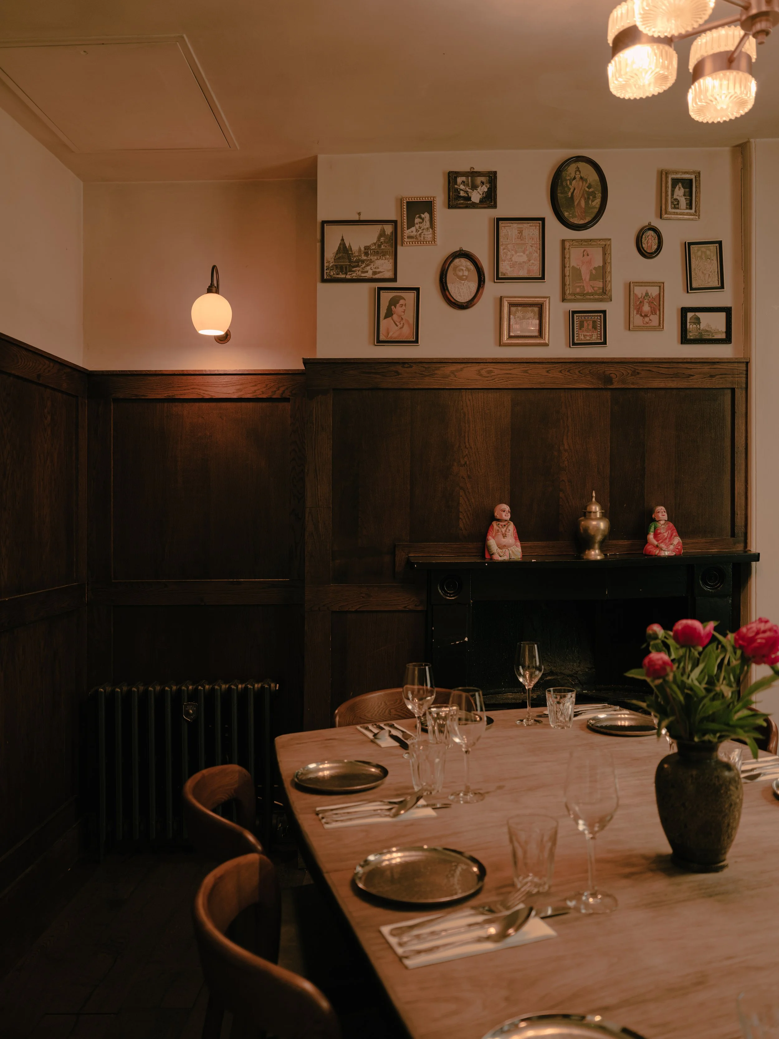A dining table set with glassware, silverware, and napkins in a warmly lit restaurant with framed artwork on the wall and decorative dolls on the mantelpiece, with a vase of pink flowers in the foreground.