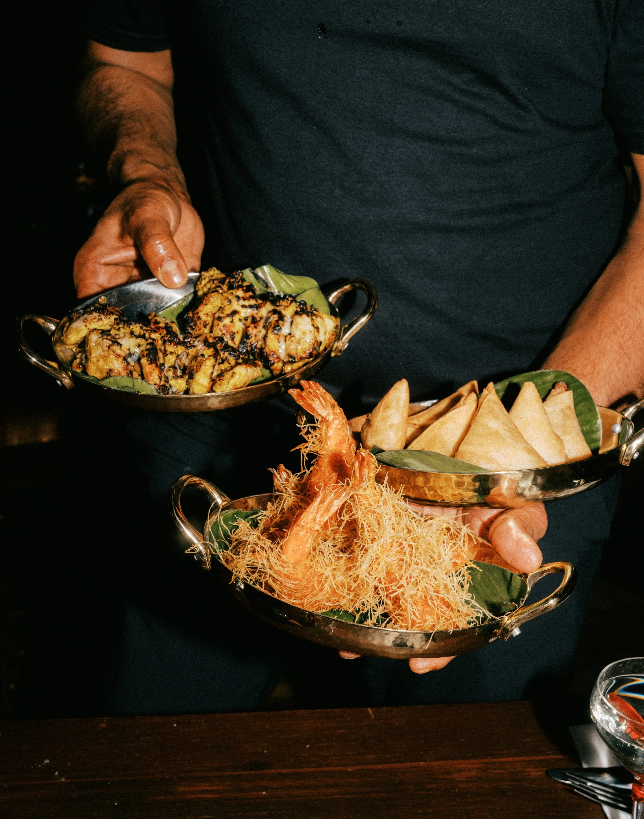 Person holding a stainless steel serving tray with grilled food, possibly chicken, garnished with black sesame seeds served on a banana leaf. Another platter with fried, cone-shaped snacks on a banana leaf is visible in the foreground.