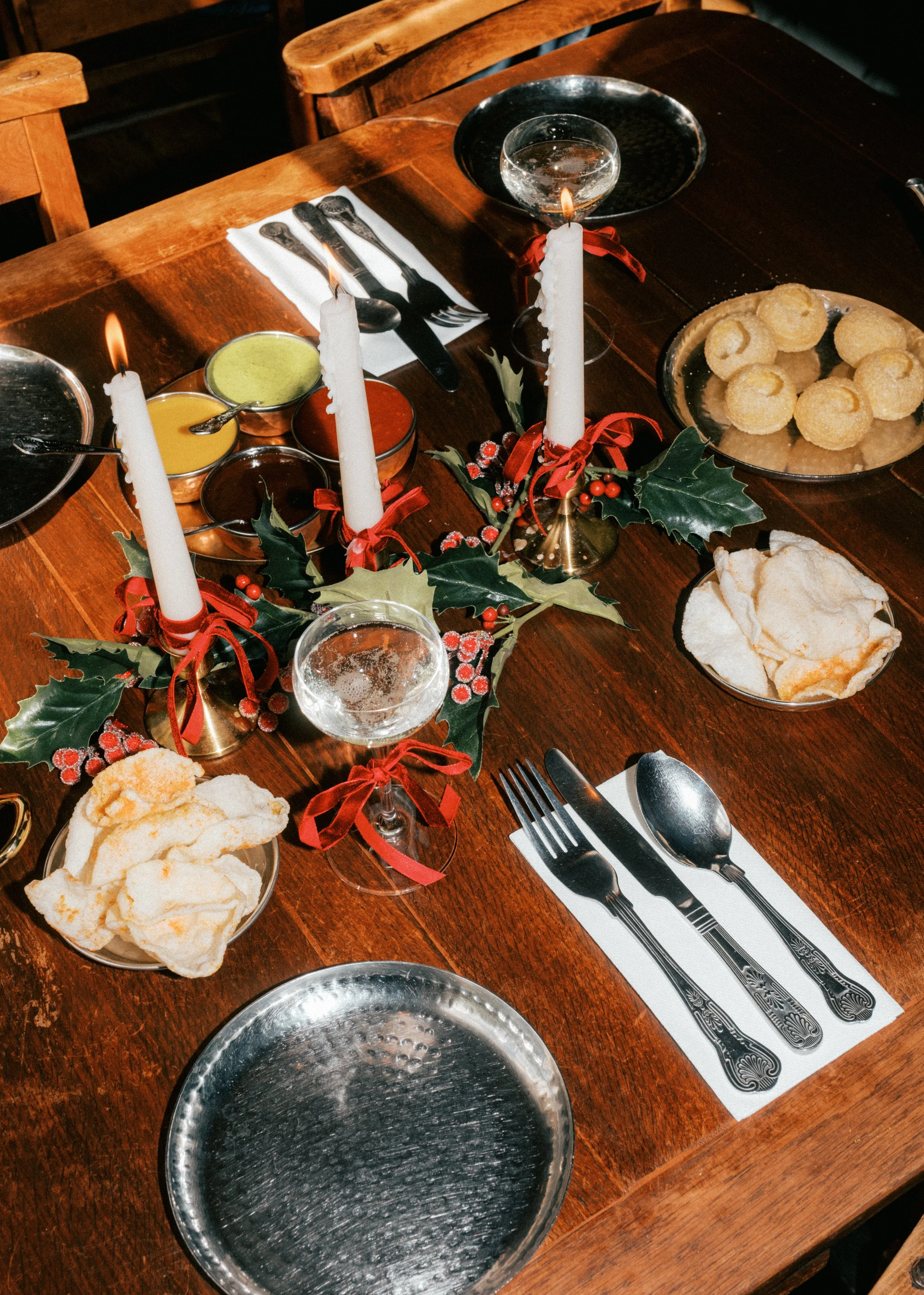 A wooden dining table set for a Christmas celebration with three lit white candles decorated with holly and red ribbons, a plate of cheese puffs, a bowl of potato chips, various sauces, and empty silver dishes, with cutlery and napkins.