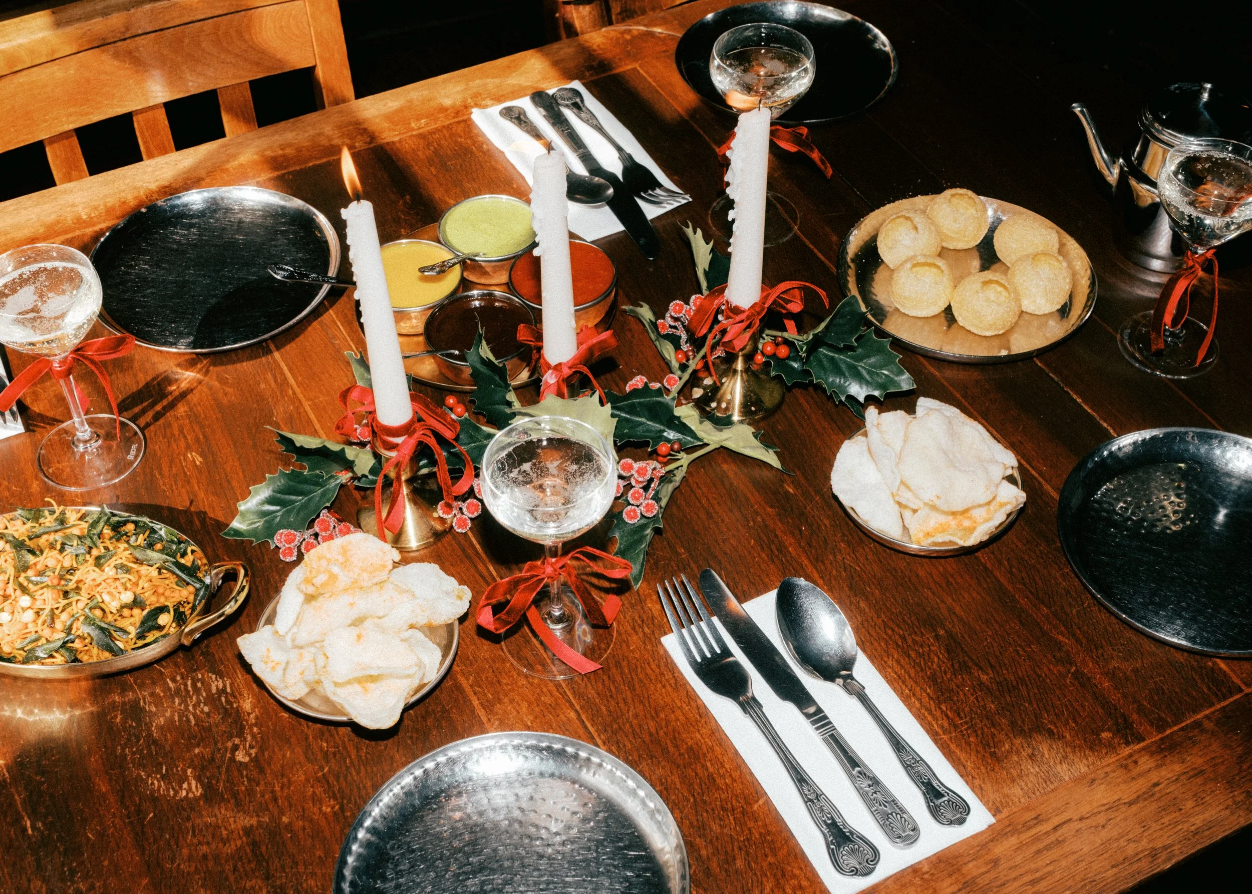 A festive holiday table setting with lit candles, holly decorations, and various dishes including a bowl of pasta with greens, fried cheese balls, and bread chips. Place settings include silverware on a white napkin with a black and white pattern.