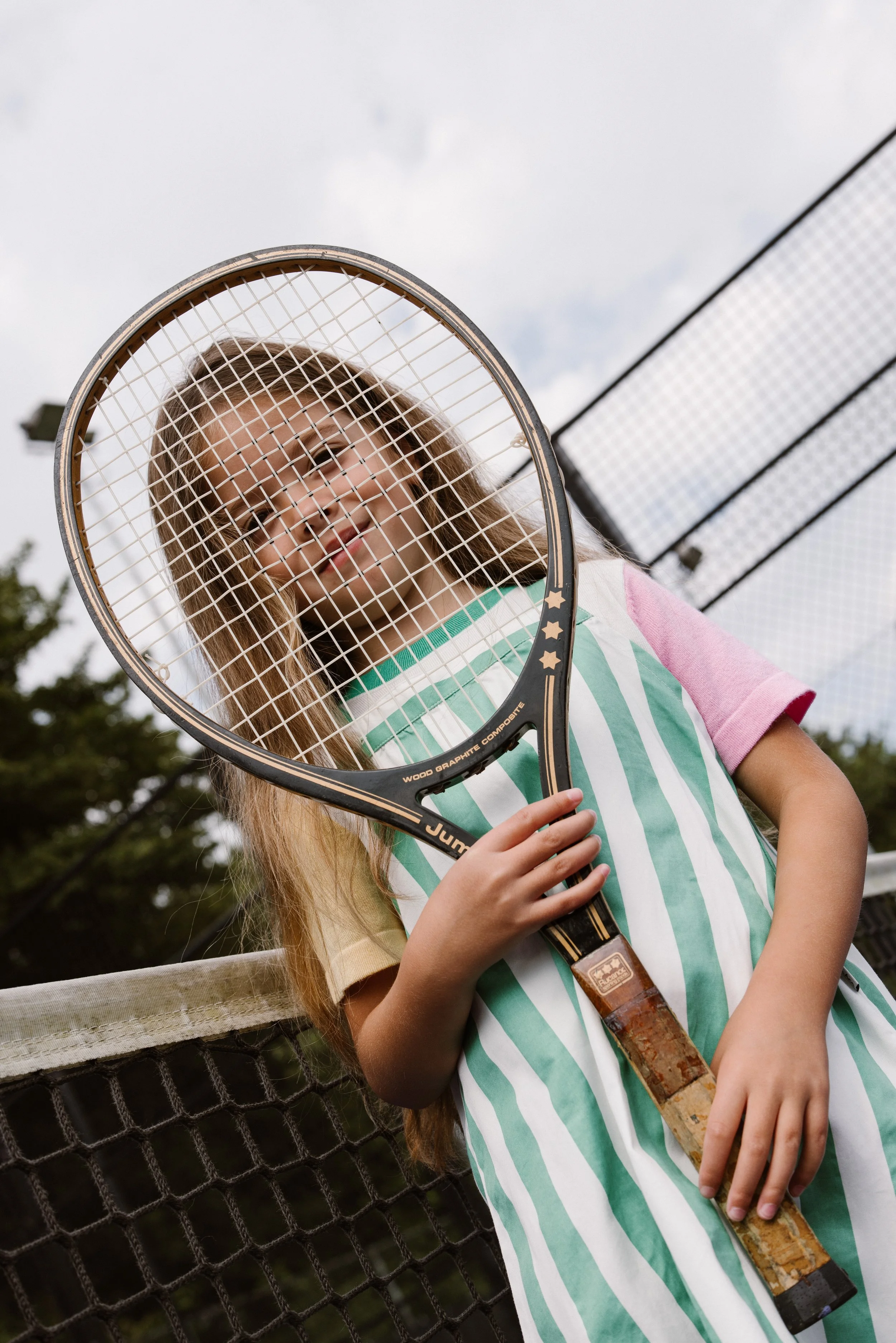 A young girl with long light brown hair holding a tennis racquet standing on a tennis court, with a chain-link fence and cloudy sky in the background. fashion shoot, lifestyle shoot, brandshoot, lieneloe fashion haarlem