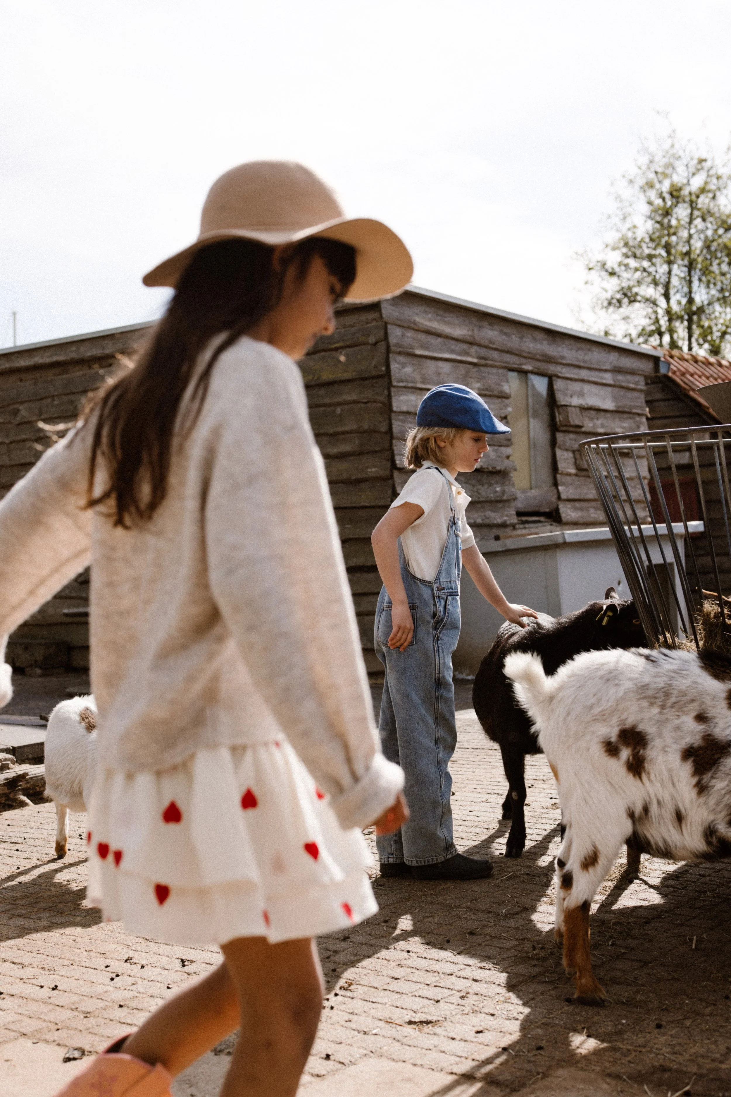 Children petting goats in a farmyard, with a girl in a white dress with red hearts and a girl in denim overalls and a blue hat, in front of a wooden shed. fashion shoot, lifestyle shoot, brandshoot, lieneloe fashion haarlem