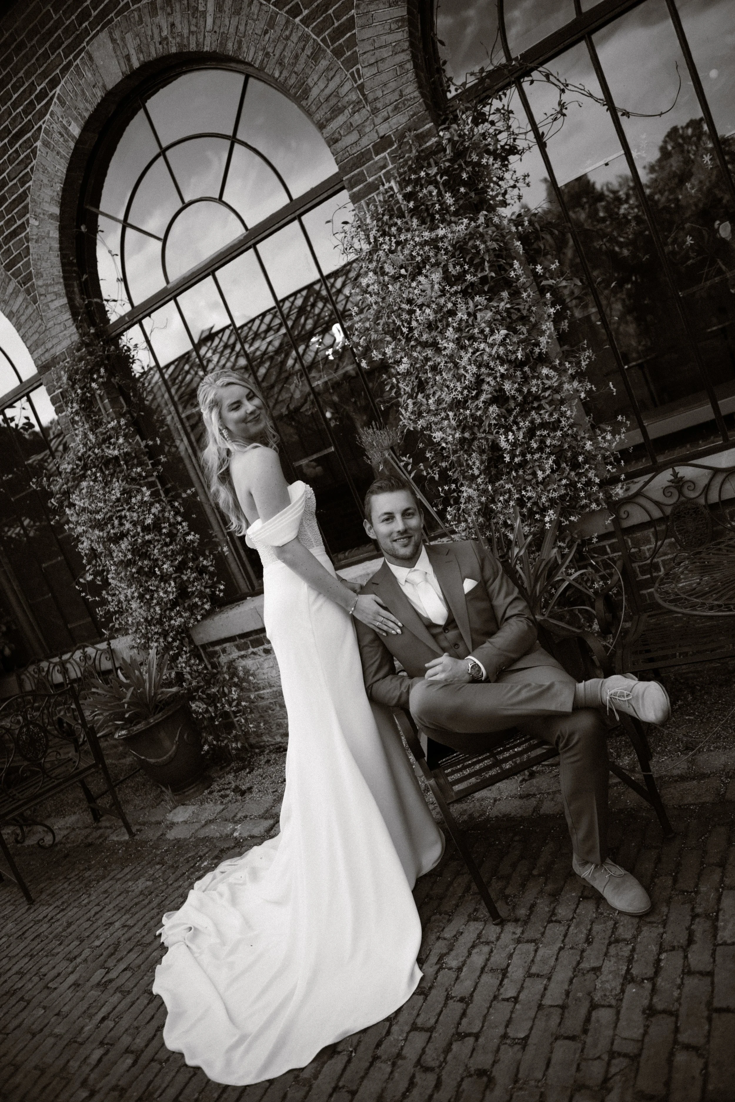 Black and white photo of a couple in formal attire, posing outdoors in front of a brick building with large arched windows and plants. The woman is standing in a white dress, and the man is sitting on a bench in a suit.
Bruidsfotograaf, Bruiloft
