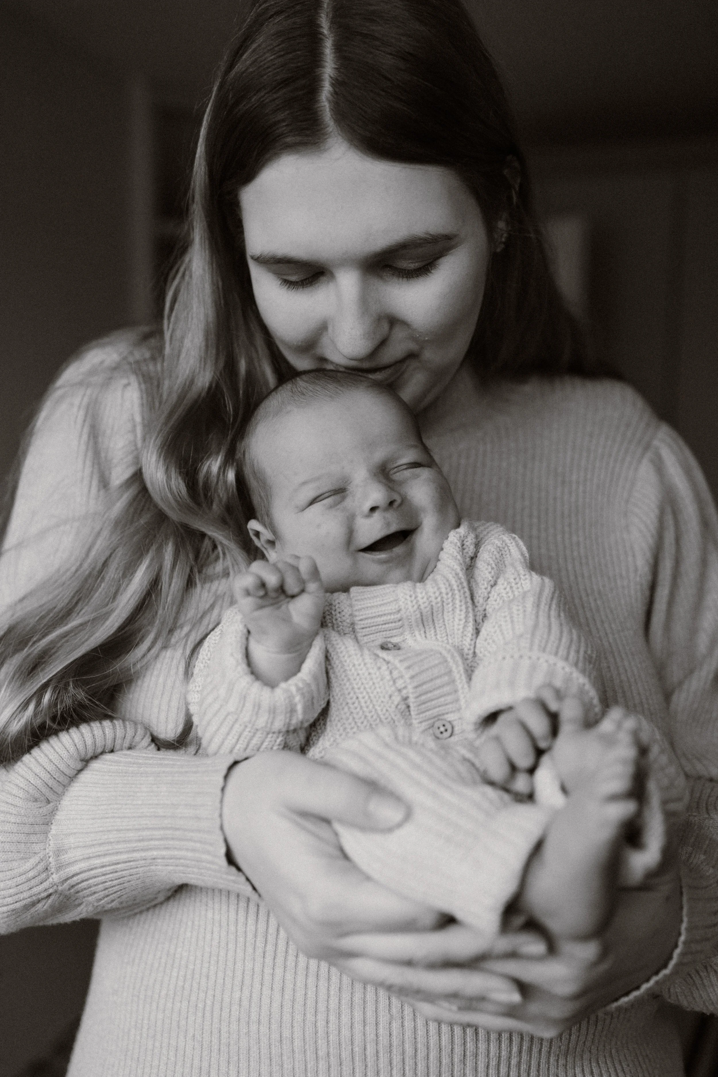 A woman holding a smiling baby in her arms, both dressed in cozy, light-colored sweaters, in a warm indoor setting. newbornshoot, newbornfotograaf haarlem, newborn shoot thuis. 