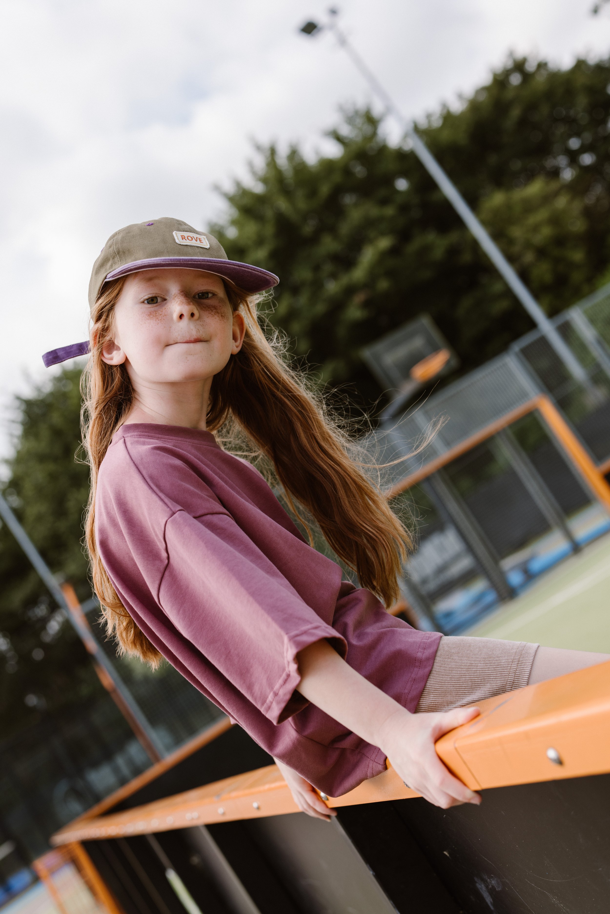 A young girl with red hair, wearing a beige cap with a purple brim and a pinkish-brown shirt, is holding onto an orange hockey stick on an outdoor hockey rink. fashion shoot, lifestyle shoot, brandshoot, lieneloe fashion haarlem