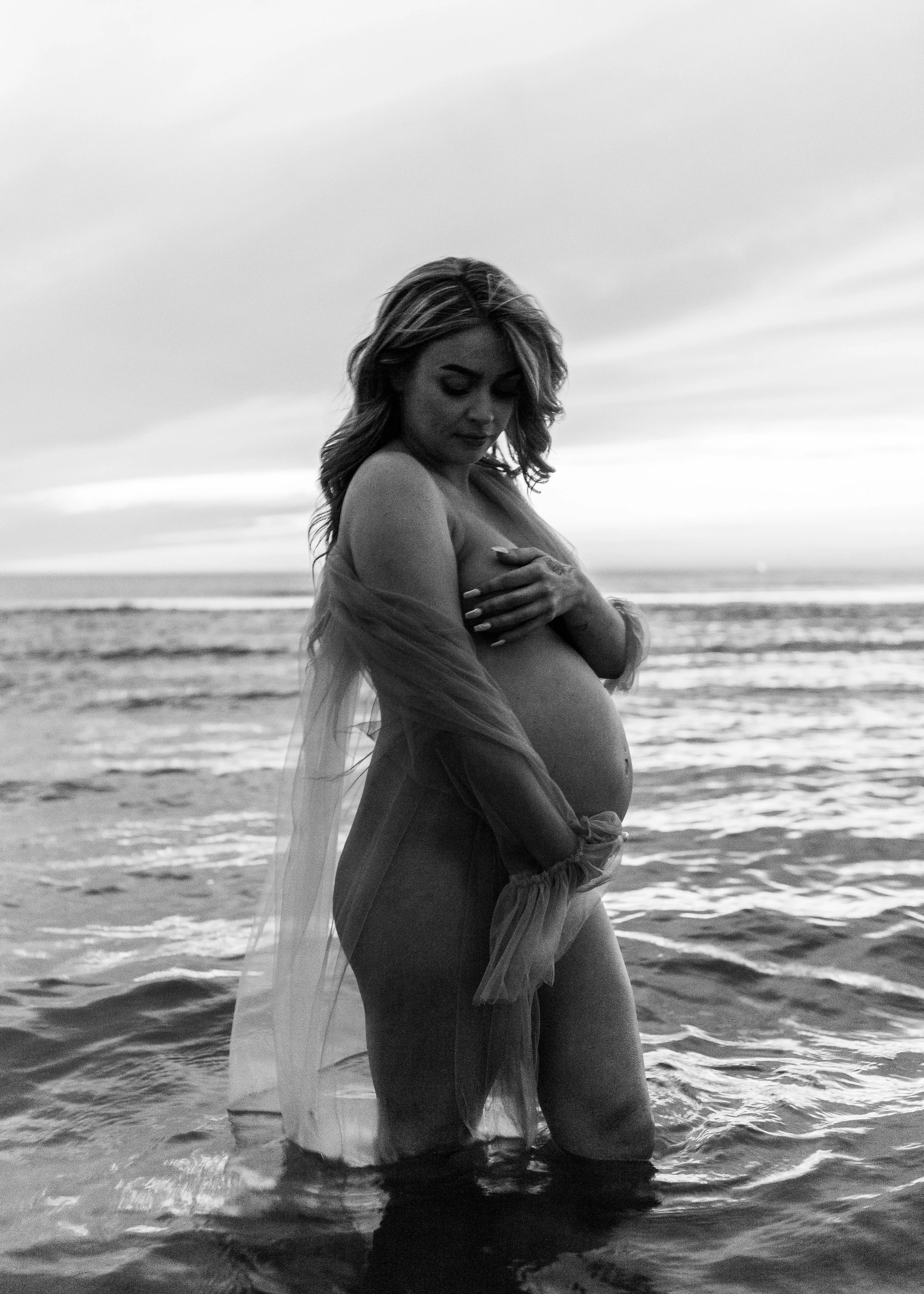 A pregnant woman wearing a white dress standing outdoors at sunset, bij ijmuiden aan zee, ijmuiderslag. lifestylefotografie, lifestyleshoot, zwangerschapsfotografie, zwangerschapsshoot, shoot duinen, fotograaf haarlem. 