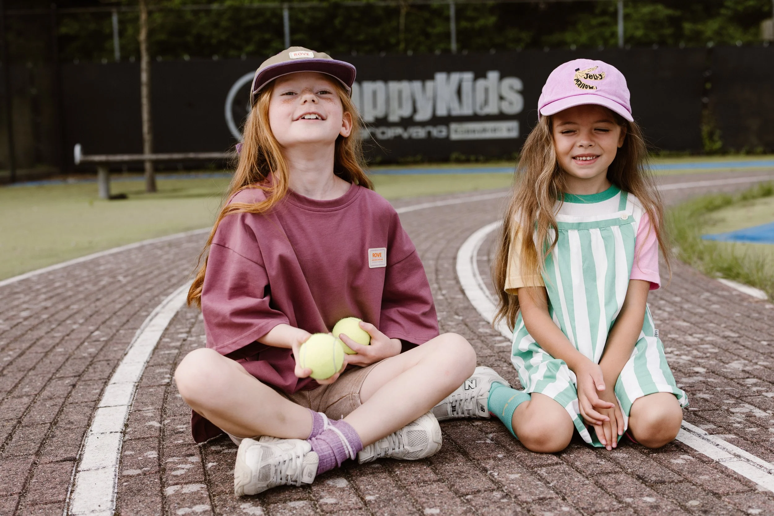 Two young girls sitting on a brick tennis court with tennis balls, wearing casual clothes and baseball caps, smiling and relaxing after playing tennis. fashion shoot, lifestyle shoot, brandshoot, lieneloe fashion haarlem