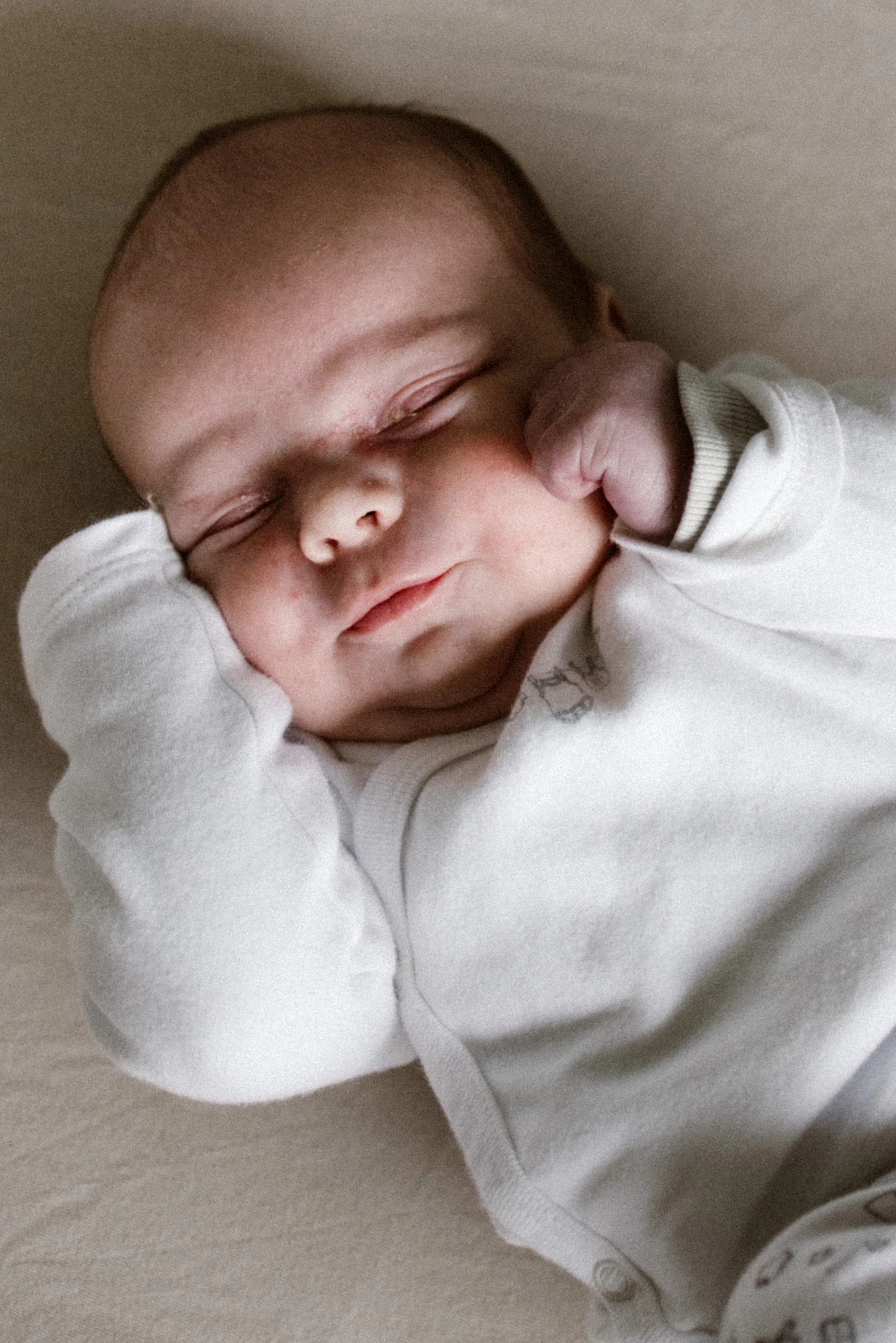 A sleeping baby with eyes closed, wearing a white long-sleeve shirt with a collar, lying on a soft surface. newbornshoot, newbornfotograaf haarlem, newborn shoot thuis. 