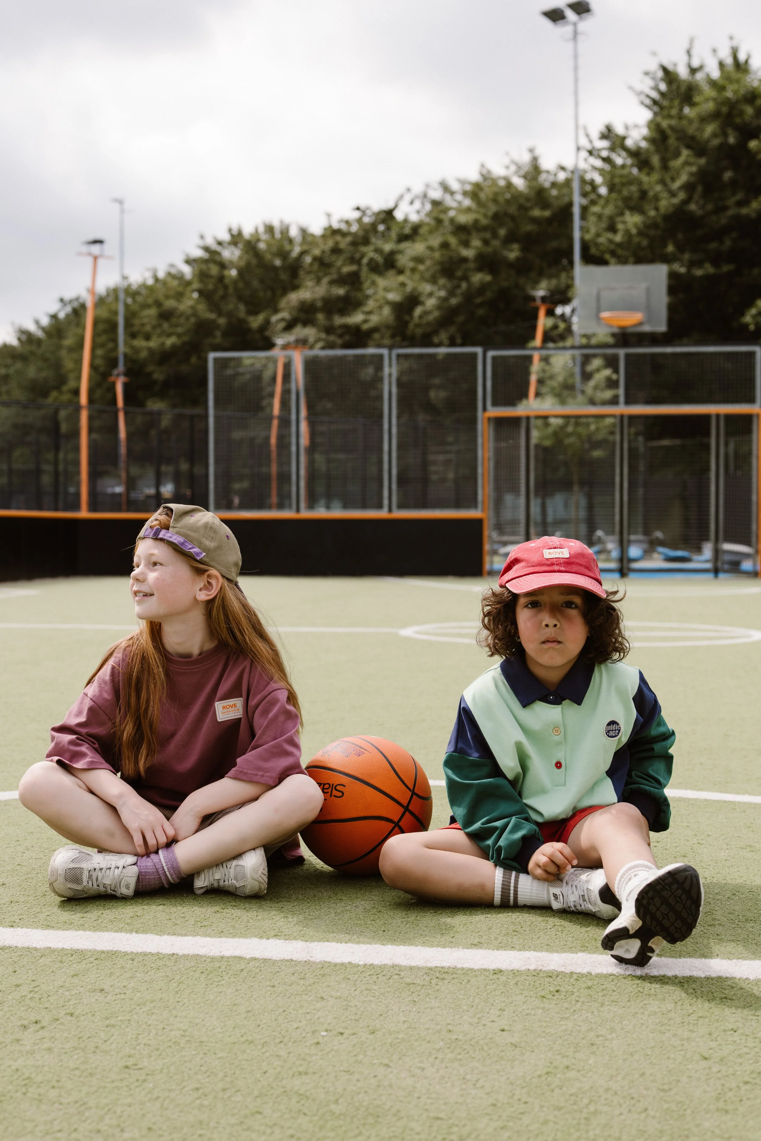 Two children sitting cross-legged on a basketball court, with a basketball ball between them. fashion shoot, lifestyle shoot, brandshoot, lieneloe fashion haarlem