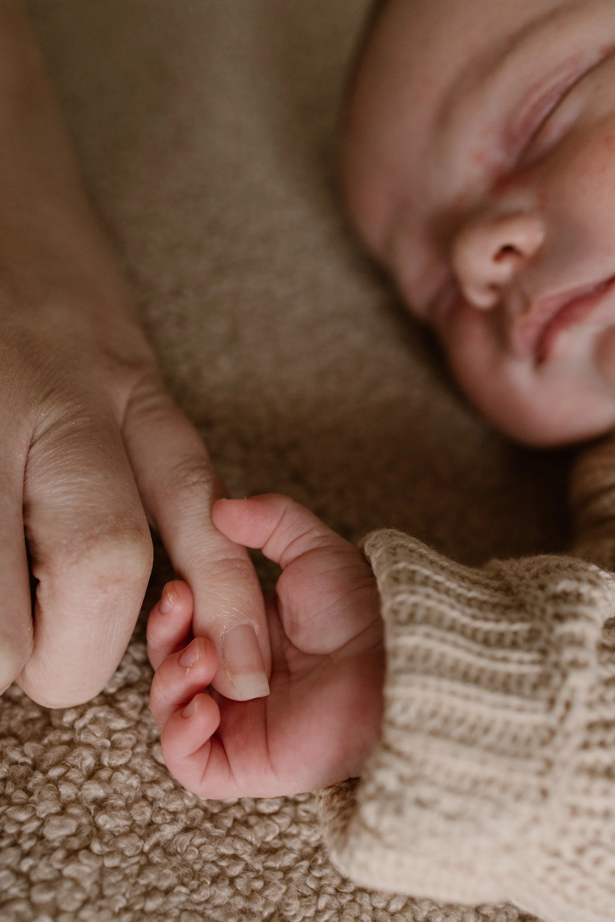 Close-up of a newborn baby's face sleeping, with a hand gently touching the baby's fingers, on a textured beige blanket. newbornshoot, newbornfotograaf haarlem, newborn shoot thuis. 