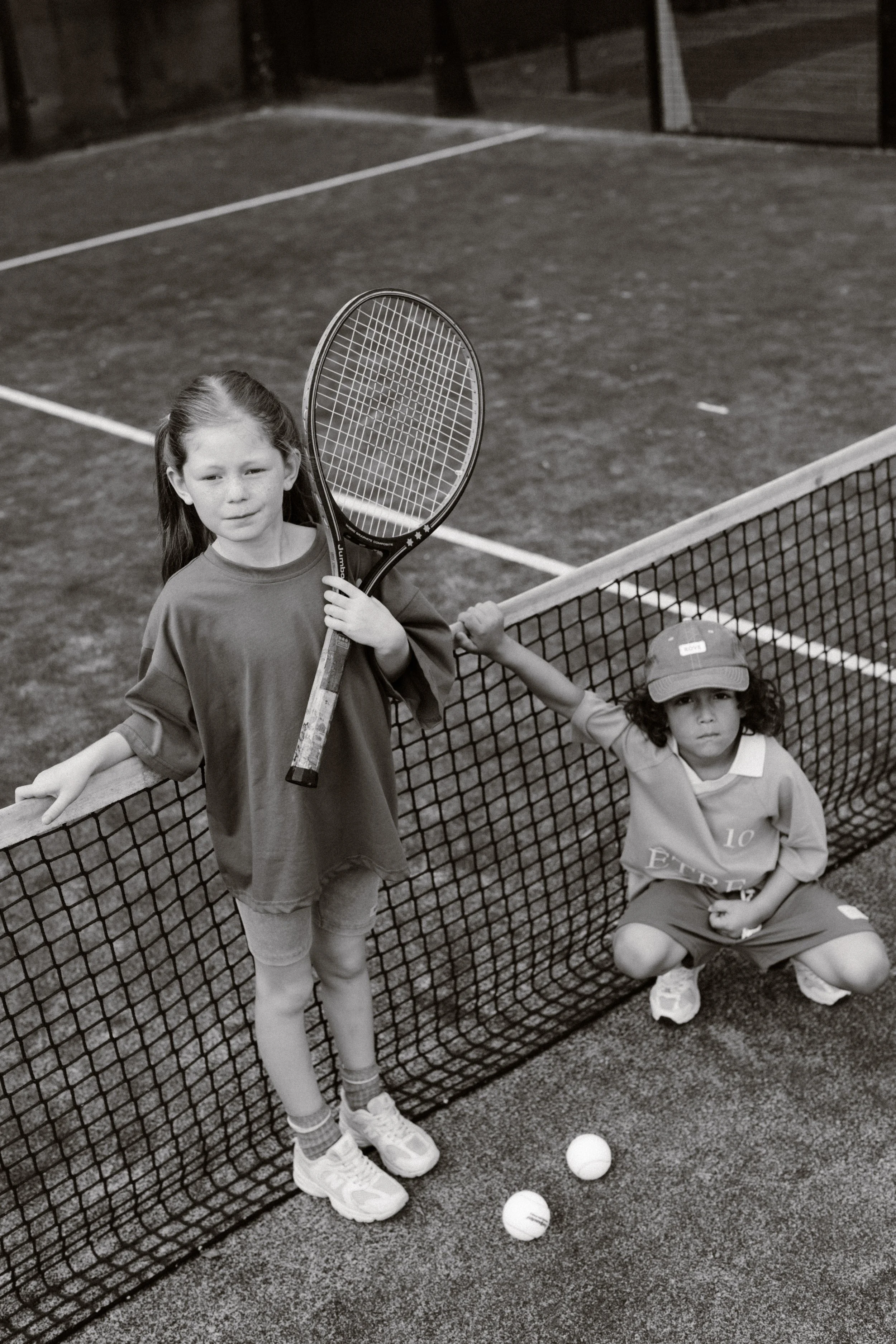 Children on a tennis court near a net, one girl standing with a tennis racket, a boy sitting on the ground with tennis balls nearby, both wearing sports clothes. fashion shoot, lifestyle shoot, brandshoot, lieneloe fashion haarlem