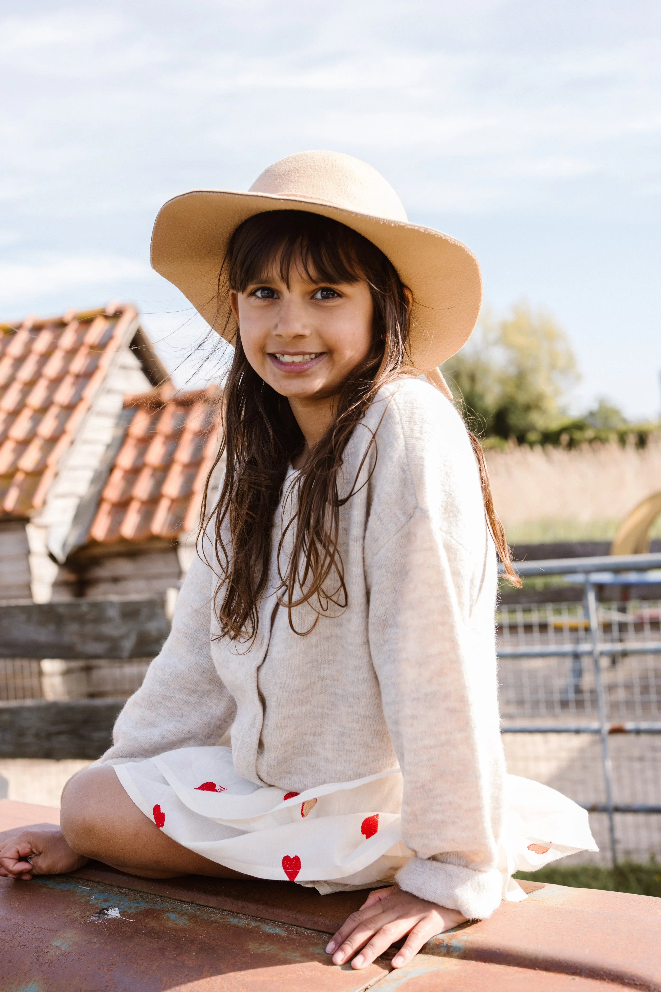 Young girl with long brown hair, wearing a wide-brimmed beige hat, cream-colored cardigan, and a white skirt with red hearts, sitting outdoors on a ledge with a clear sky. fashion shoot, lifestyle shoot, brandshoot, lieneloe fashion haarlem
