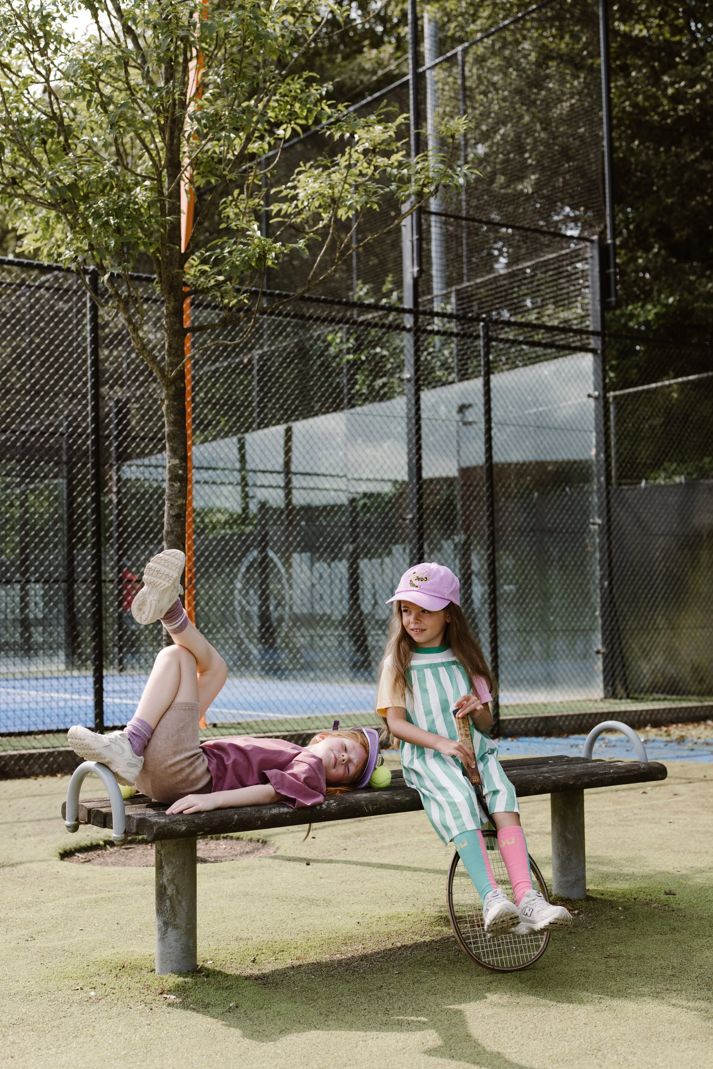 Three young girls playing at a park near a tennis court. One girl in a maroon shirt and beige shorts is lying on a bench with her feet up in the air. fashion shoot, lifestyle shoot, brandshoot, lieneloe fashion haarlem
