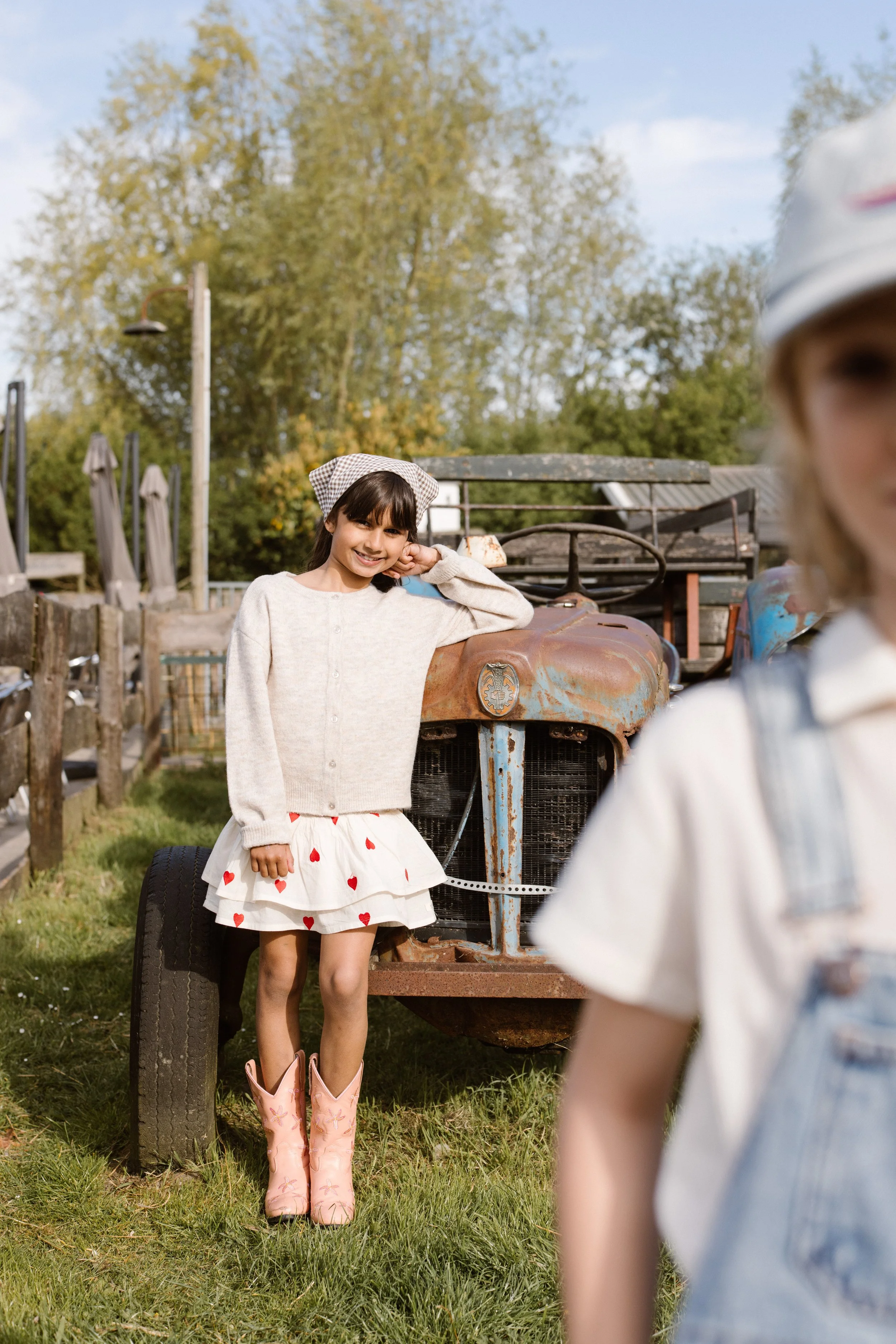 A girl with dark hair smiling and leaning on an old rusty tractor, wearing a light-colored sweater and a white skirt with red hearts, pink cowboy boots, and a checked headscarf. fashion shoot, lifestyle shoot, brandshoot, lieneloe fashion haarlem