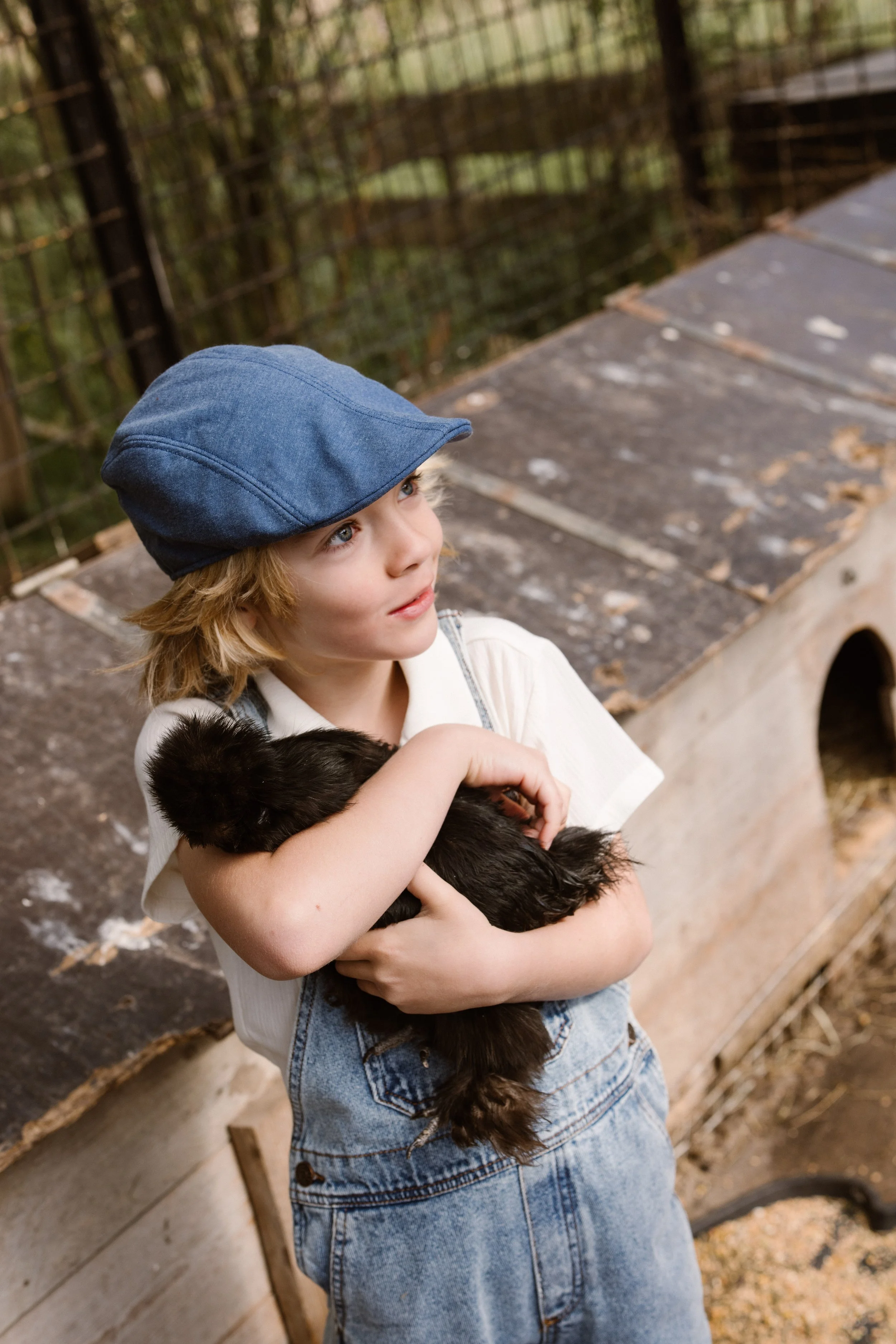 A child with blonde hair is holding a chicken in his arms at a petting zoo or farm, with a wooden and wire fence behind him. fashion shoot, lifestyle shoot, brandshoot, lieneloe fashion haarlem