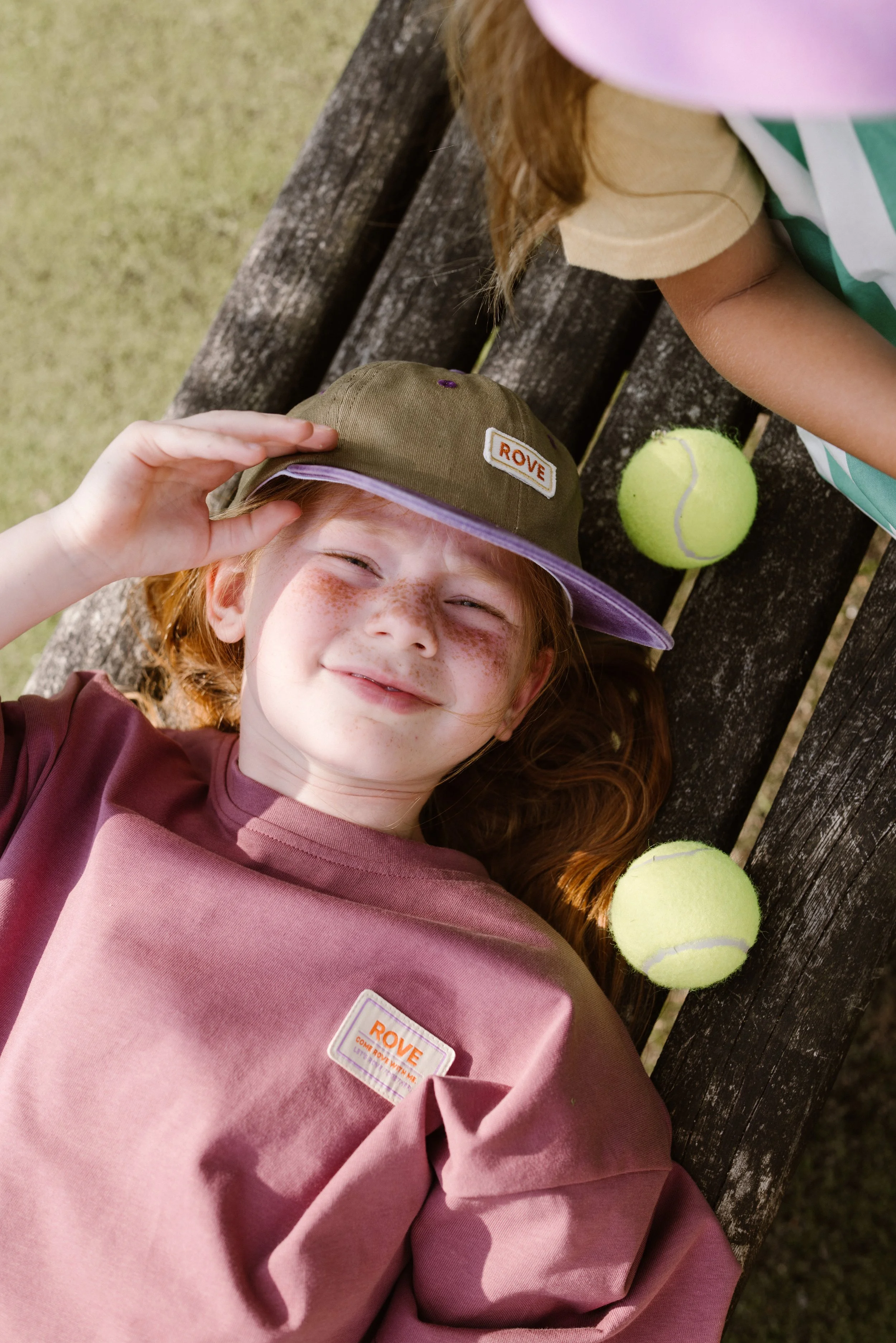 A young girl with red hair and freckles lying on a wooden bench with tennis balls beside her, wearing a beige hat and a pink sweatshirt. A fashion shoot, lifestyle shoot, brandshoot, lieneloe fashion haarlem