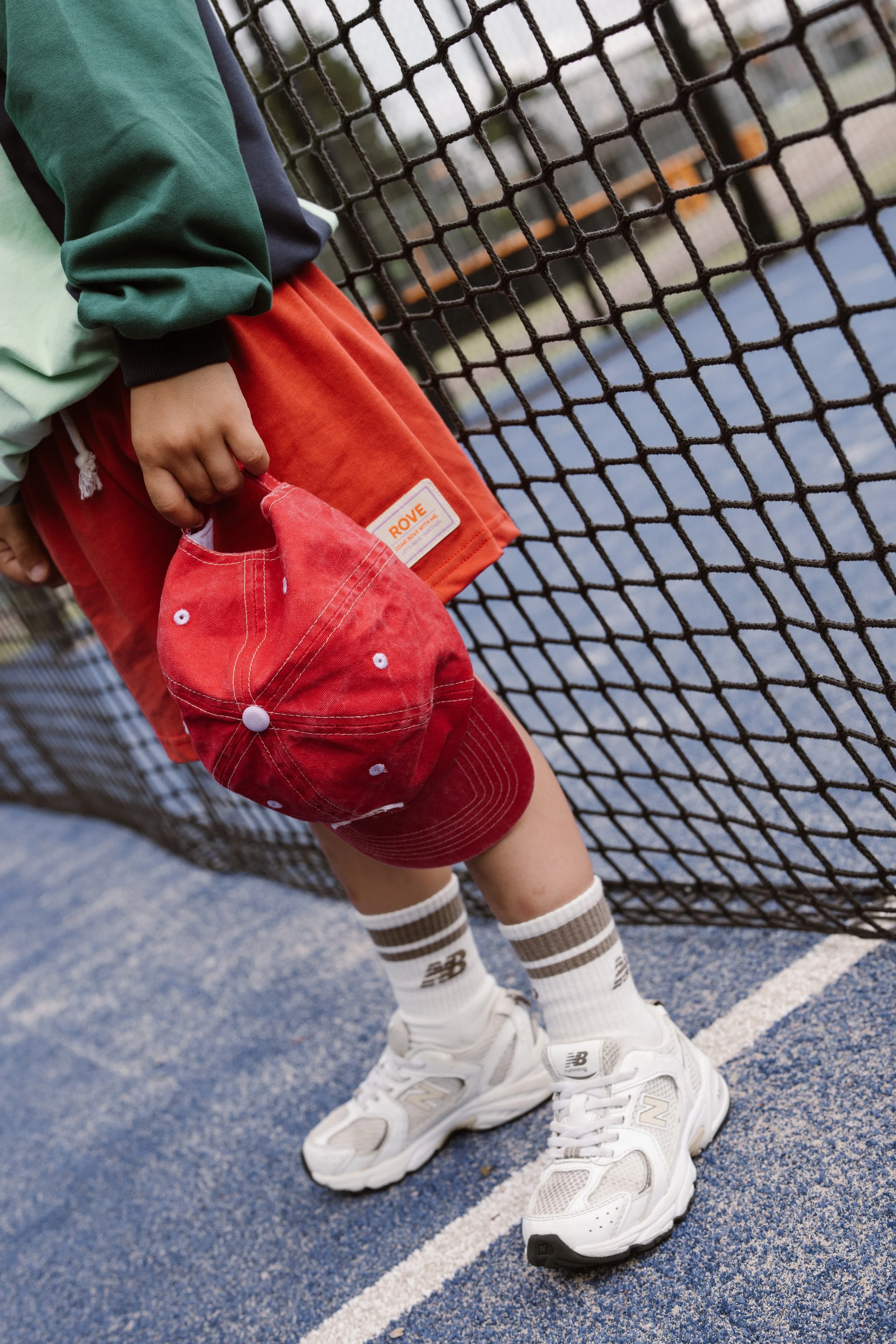 Child wearing white sneakers with black and grey accents, brown and white striped socks, red shorts with a ROVE label, green and navy jacket. fashion shoot, lifestyle shoot, brandshoot, lieneloe fashion haarlem