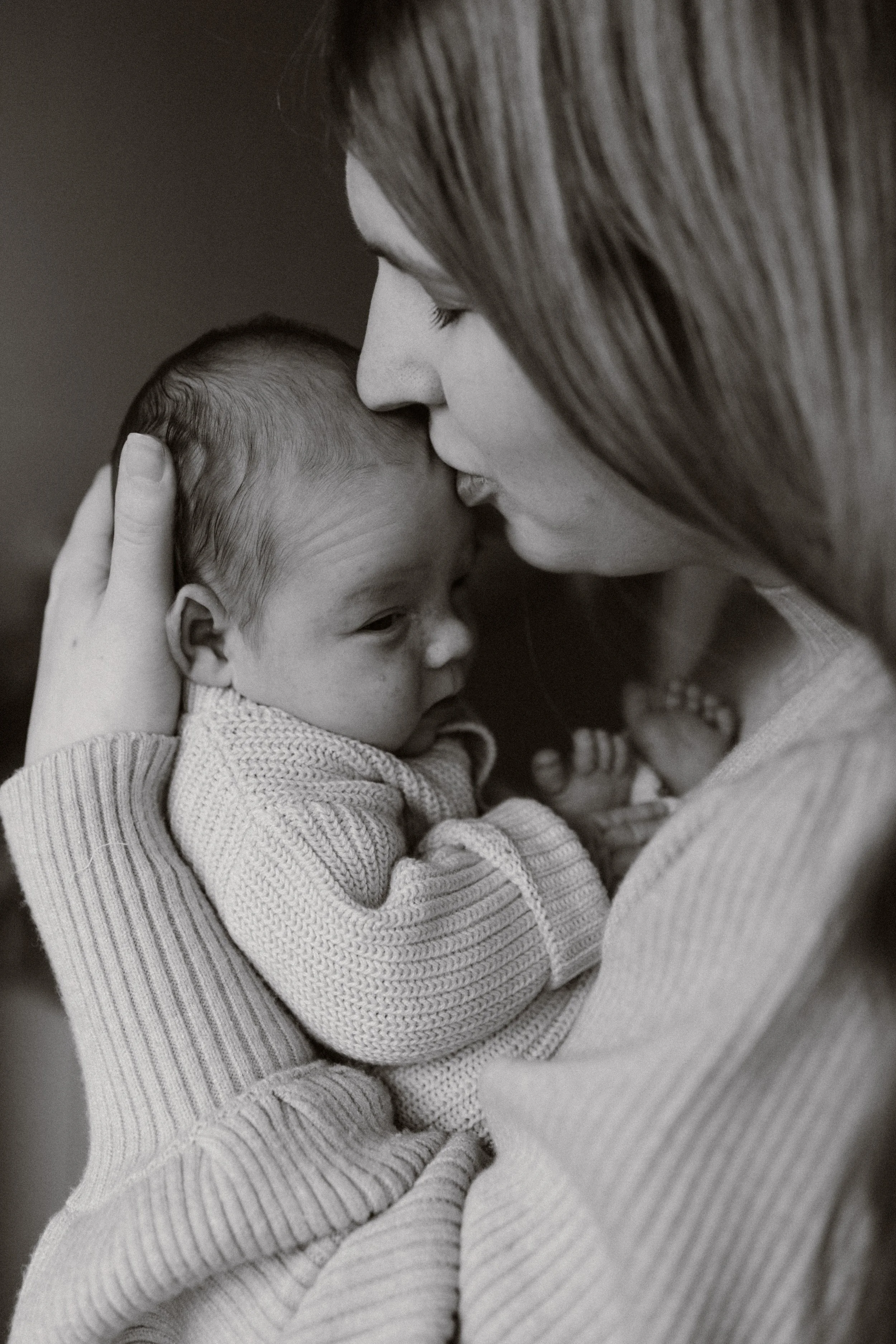 A woman lovingly holding and kissing her newborn baby, both dressed in cozy sweaters, in a tender moment of affection. newbornshoot, newbornfotograaf haarlem, newborn shoot thuis. 