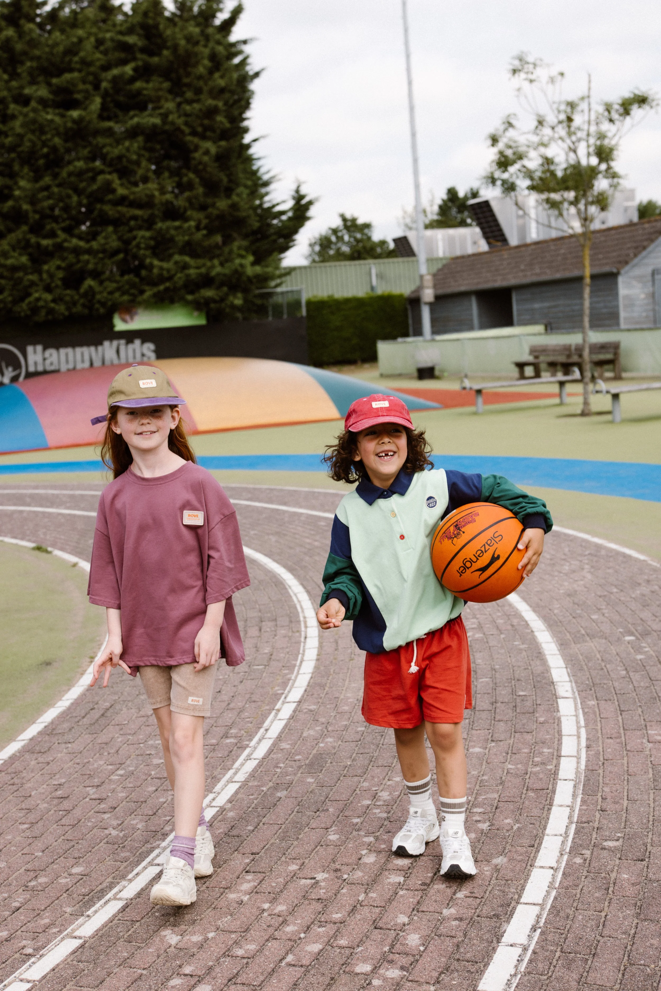 Two young kids, a girl with long red hair wearing a hat and a boy with curly hair wearing a red cap, are playing basketball on a track field. fashion shoot, lifestyle shoot, brandshoot, lieneloe fashion haarlem