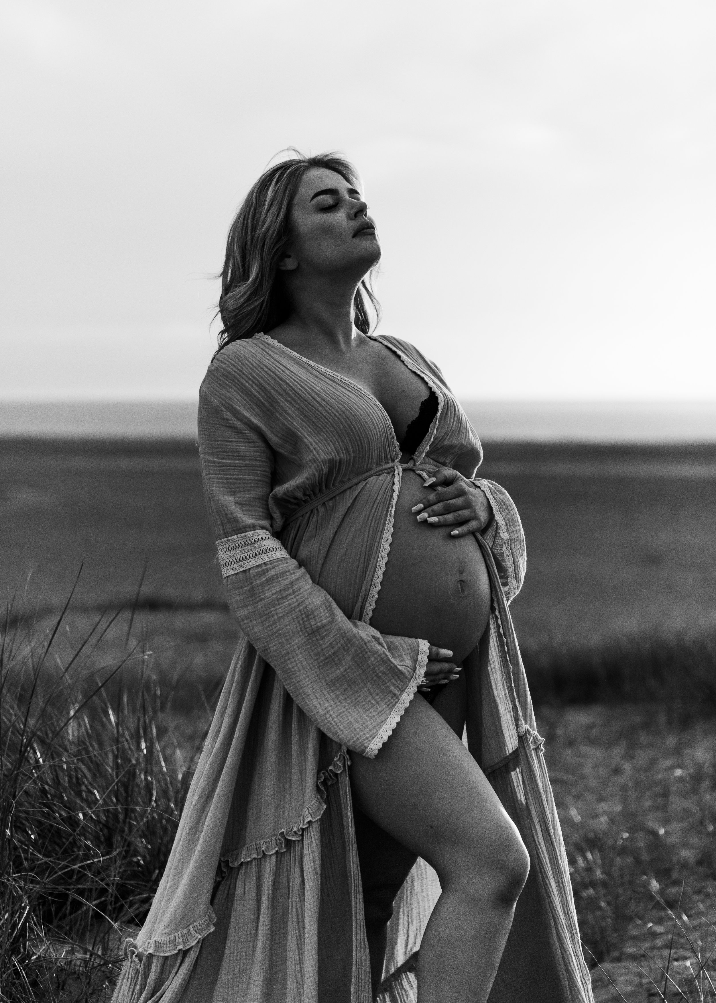 A pregnant woman wearing a white dress standing outdoors at sunset, bij ijmuiden aan zee, ijmuiderslag. lifestylefotografie, lifestyleshoot, zwangerschapsfotografie, zwangerschapsshoot, shoot duinen, fotograaf haarlem. 
