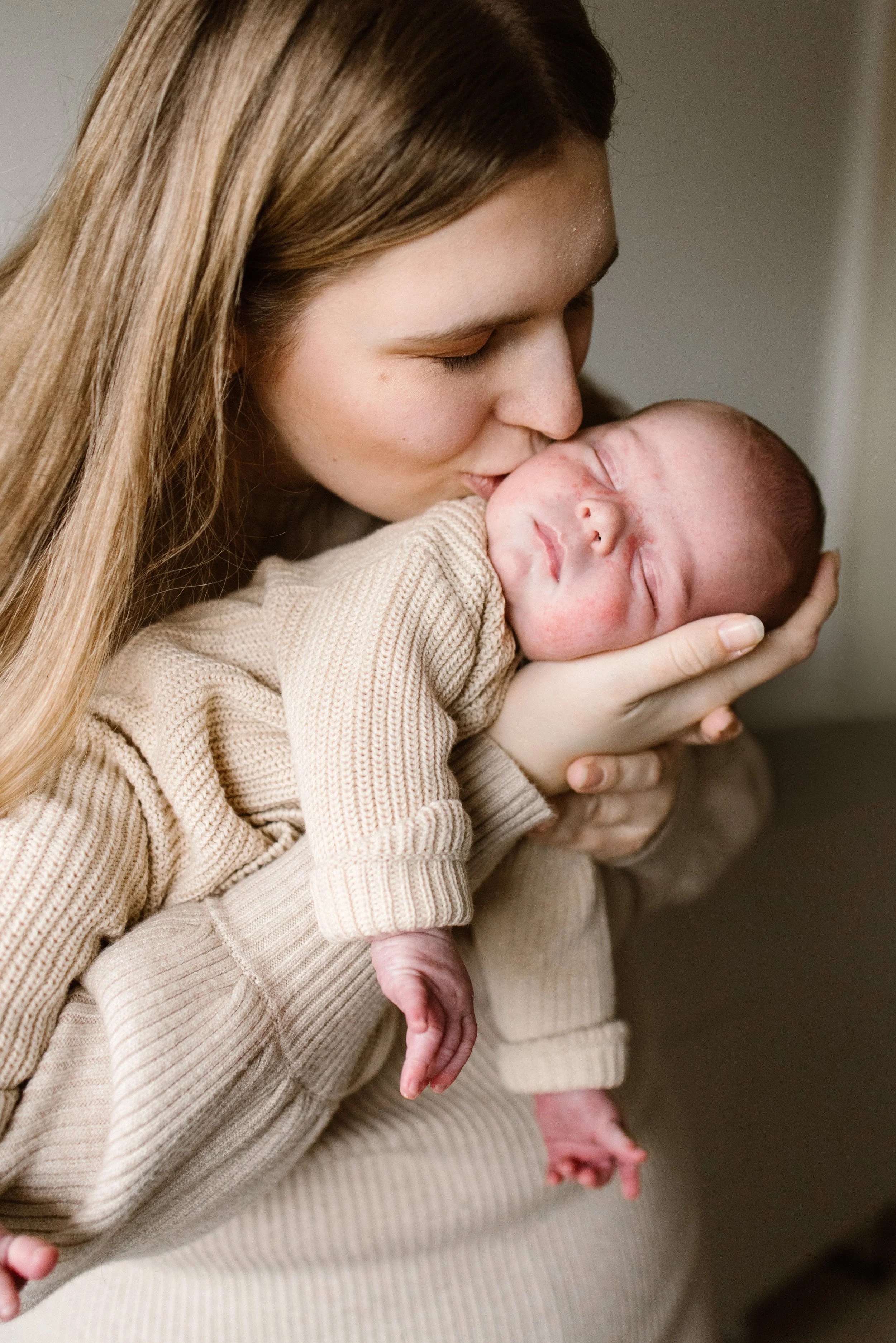A mama with long brown hair holding a sleeping newborn baby close to her face, kissing the baby's forehead.  newbornshoot, newbornfotograaf haarlem, newborn shoot thuis. 
