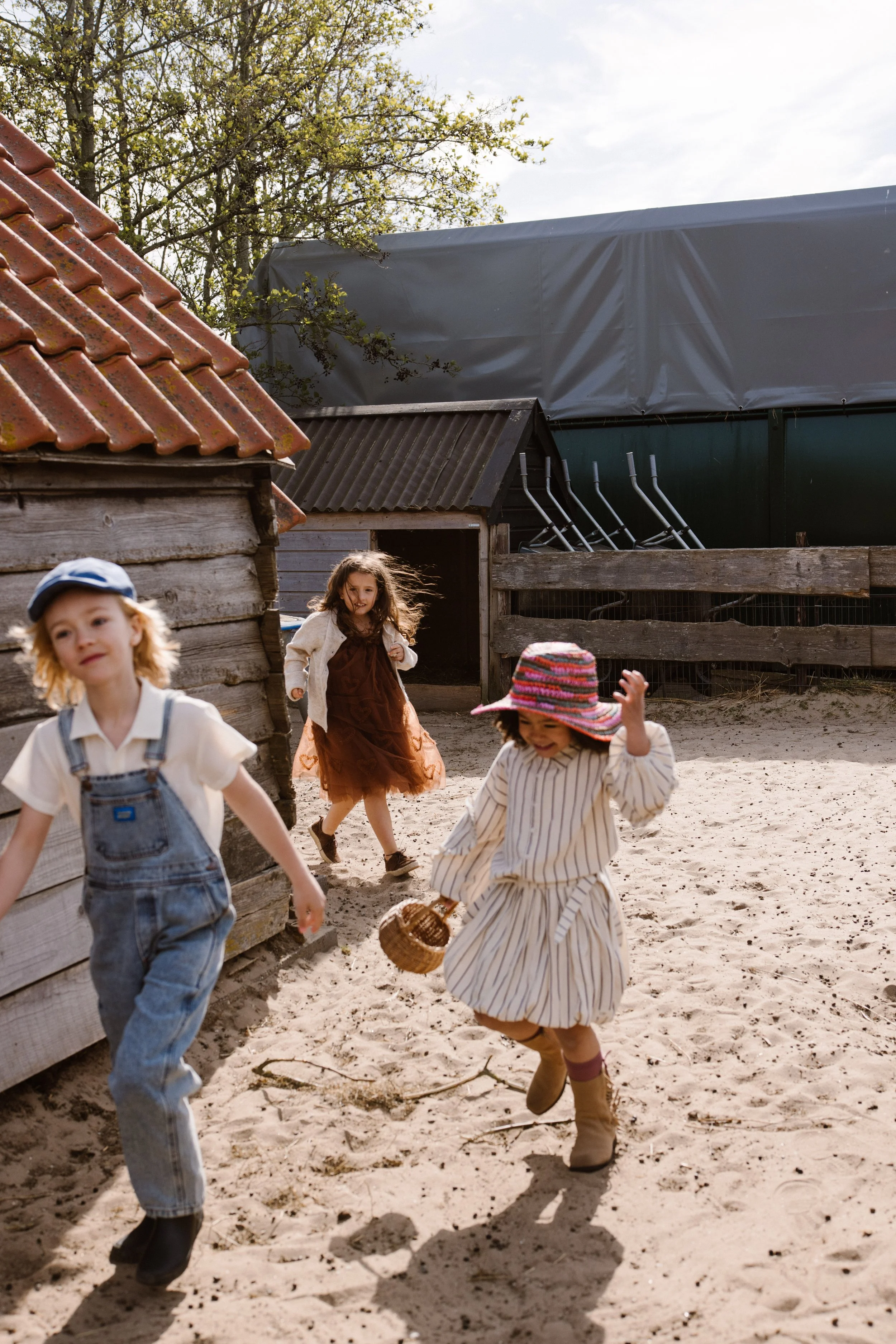 Children playing outside in a sandy area near wooden structures with a tree and a fence in the background. fashion shoot, lifestyle shoot, brandshoot, lieneloe fashion haarlem