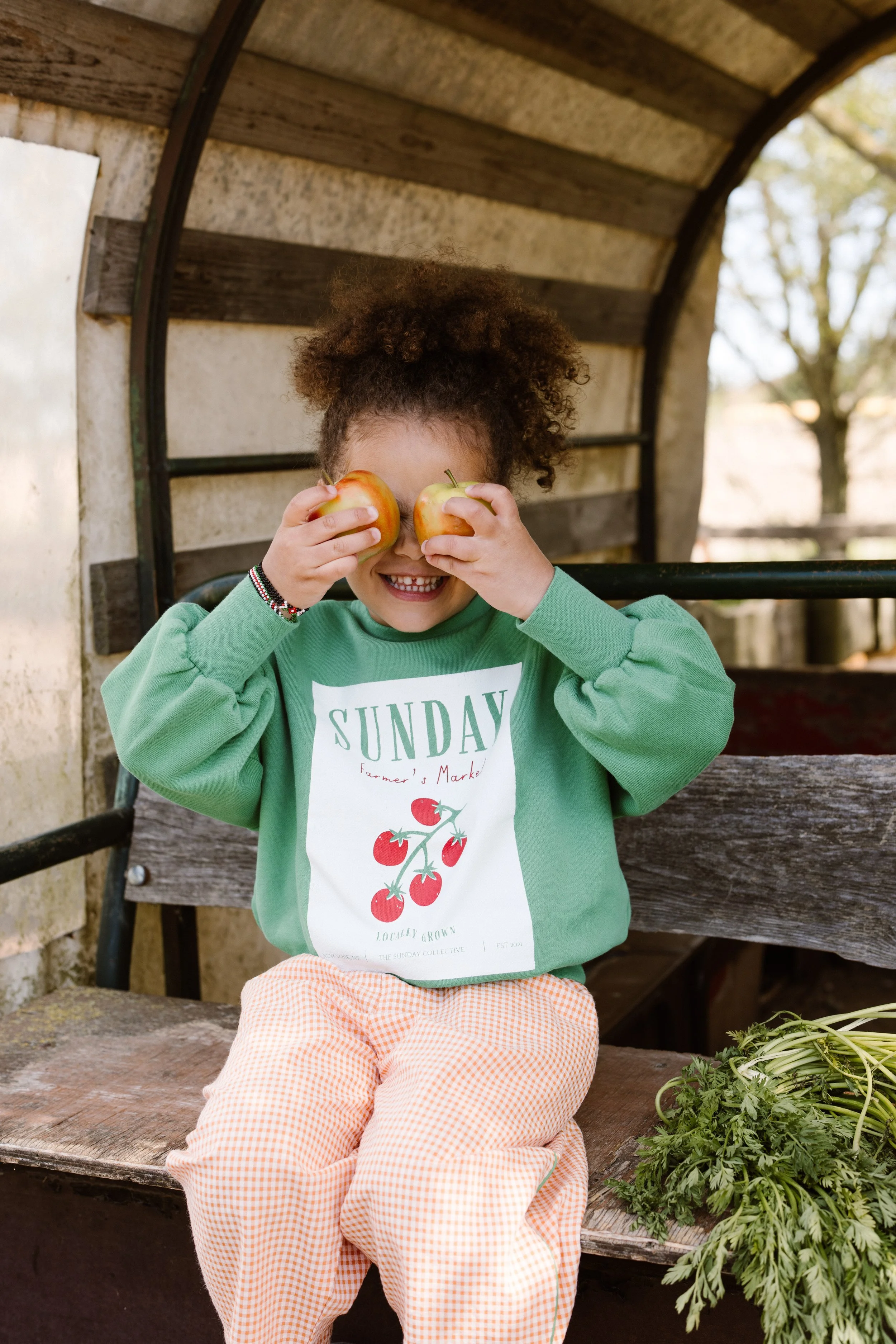 A young girl with curly hair sitting on a wooden bench, holding two apples in front of her eyes, smiling at the camera. fashion shoot, lifestyle shoot, brandshoot, lieneloe fashion haarlem