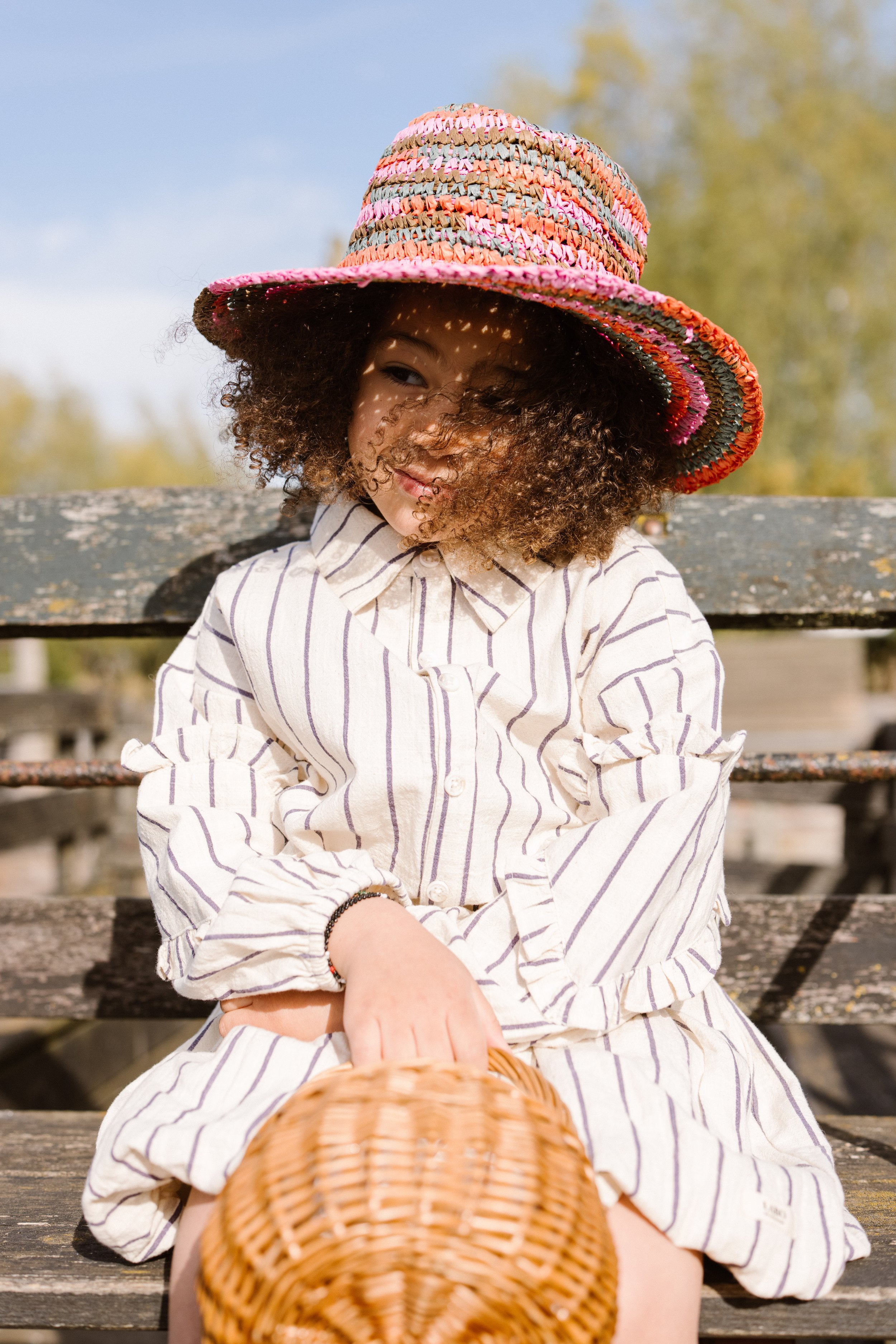 A young woman with curly hair sitting outdoors on a wooden bench, wearing a large colorful hat and a striped beige and white button-up shirt, holding a woven basket in her lap. fashion shoot, lifestyle shoot, brandshoot, lieneloe fashion haarlem