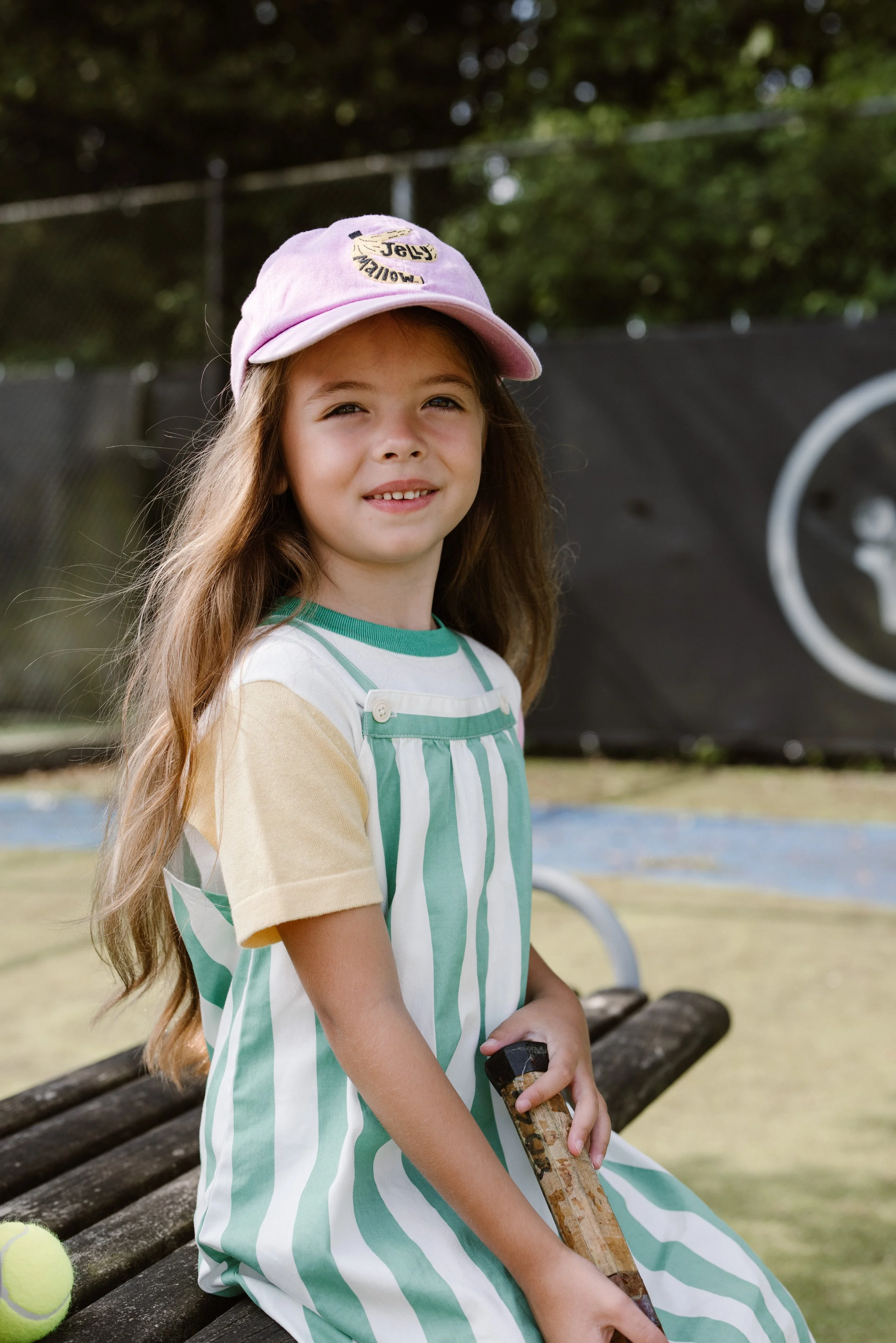 Young girl with long hair wearing a pink cap and a green and white striped dress, sitting on a bench at a tennis court, holding a tennis racket, with tennis balls nearby. fashion shoot, lifestyle shoot, brandshoot, lieneloe fashion haarlem