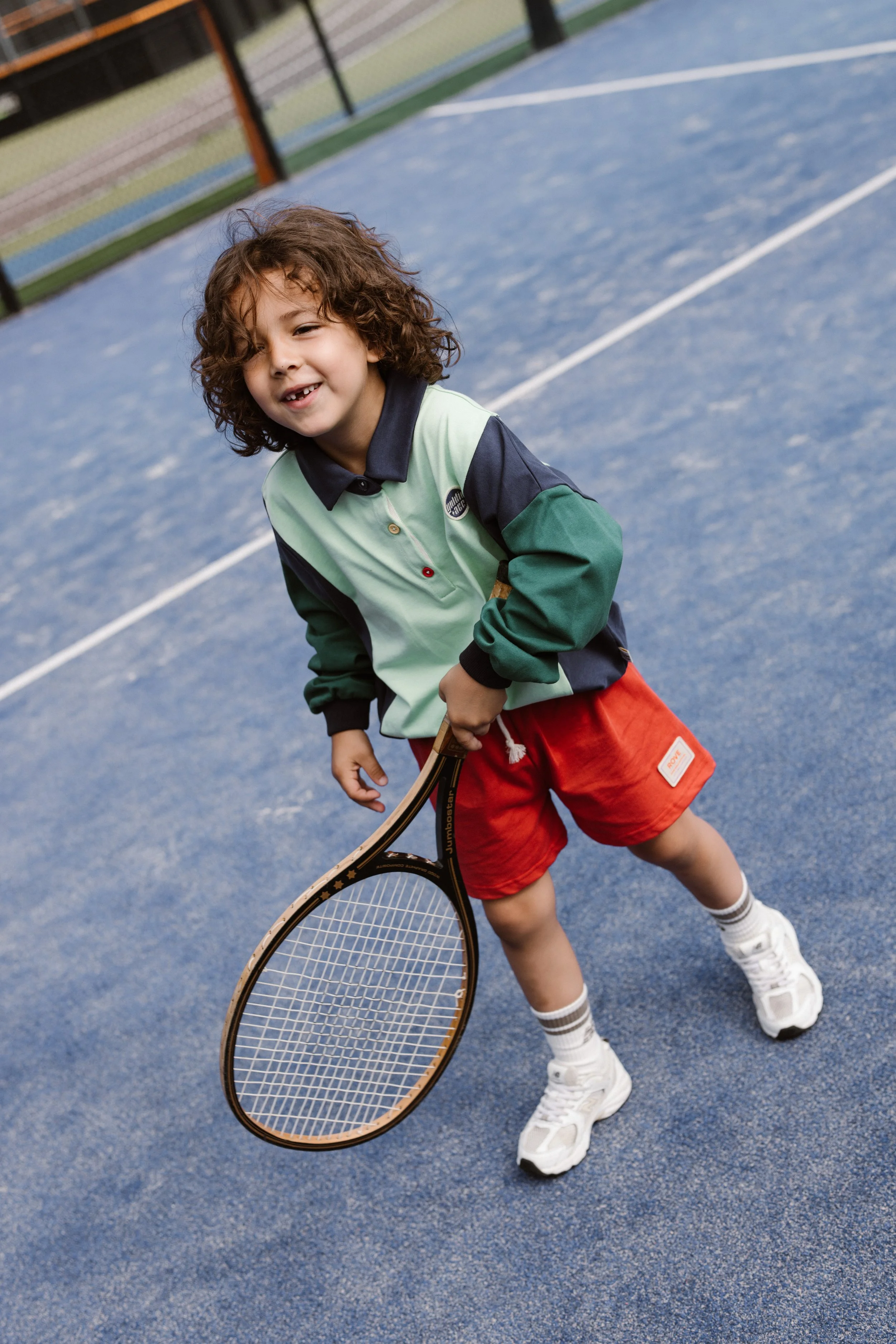 Young boy with curly brown hair playing tennis on outdoor court, wearing a light green and navy polo shirt, red shorts, white shoes, and striped socks, holding a tennis racket. fashion shoot, lifestyle shoot, brandshoot, lieneloe fashion haarlem