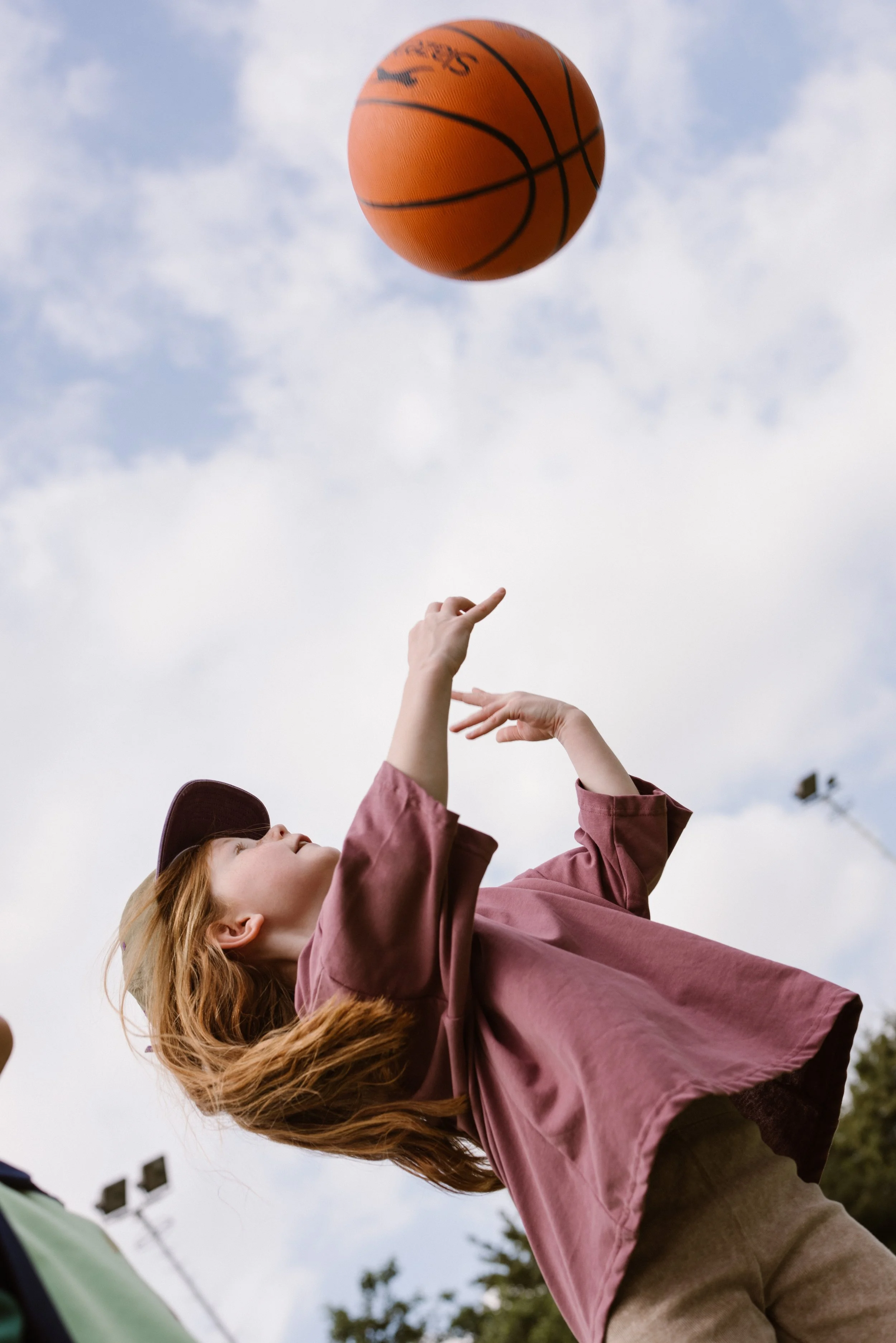 A young girl with red hair playing basketball outdoors during daytime, reaching up as the ball floats near her hands. fashion shoot, lifestyle shoot, brandshoot, lieneloe fashion haarlem