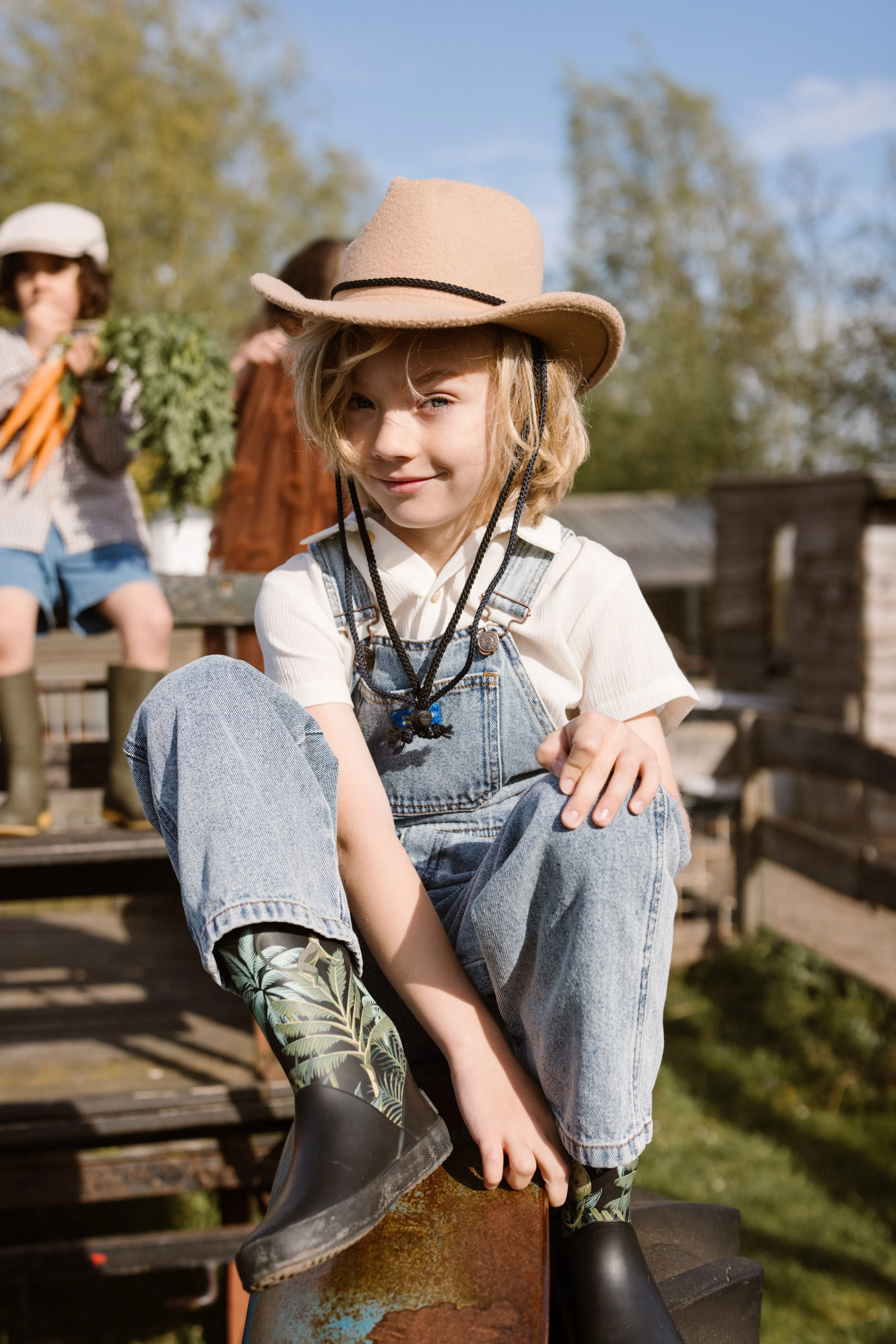 A young child with blonde hair, wearing a beige cowboy hat and overalls, sitting on a wooden surface outdoors, smiling at the camera. fashion shoot, lifestyle shoot, brandshoot, lieneloe fashion haarlem