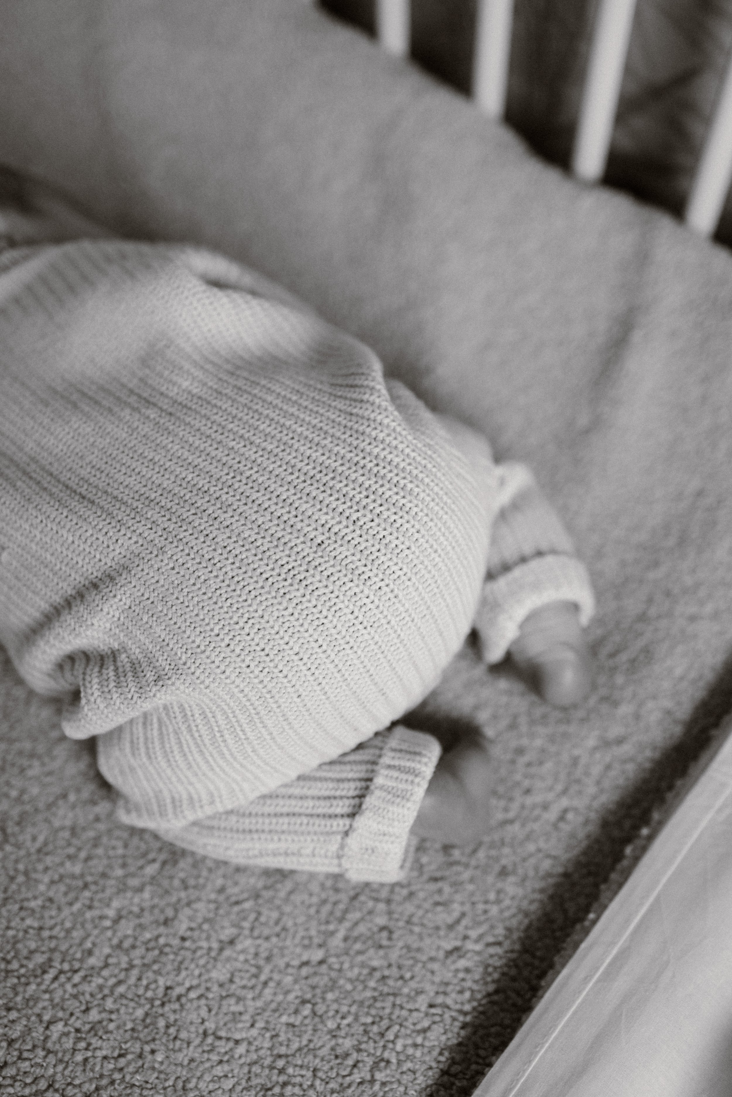 Close-up of a baby in a knitted sweater lying on a bed with blankets, with a bed rail visible in the background. newbornshoot, newbornfotograaf haarlem, newborn shoot thuis. 