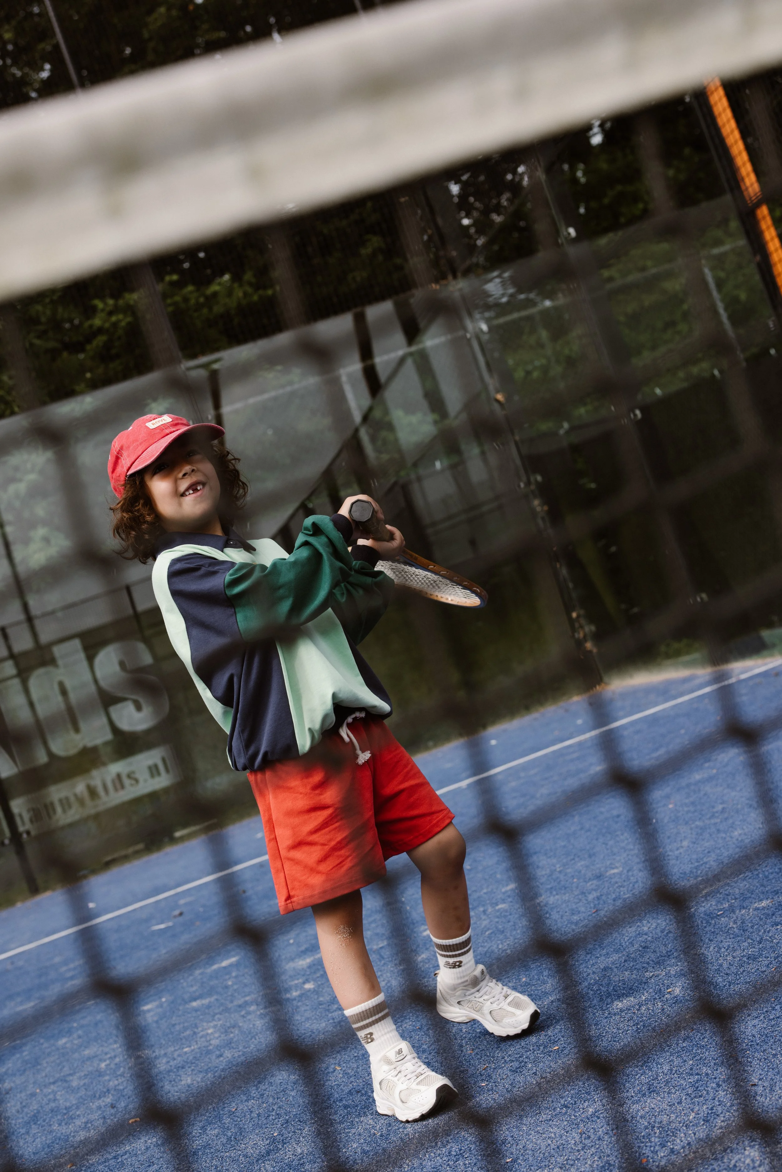 A young boy wearing a red cap, striped socks, white sneakers, and red shorts is smiling on a blue tennis court while holding a tennis racket. The image is taken through a wire fence. fashion shoot, lifestyle shoot, brandshoot, lieneloe fashion haarle