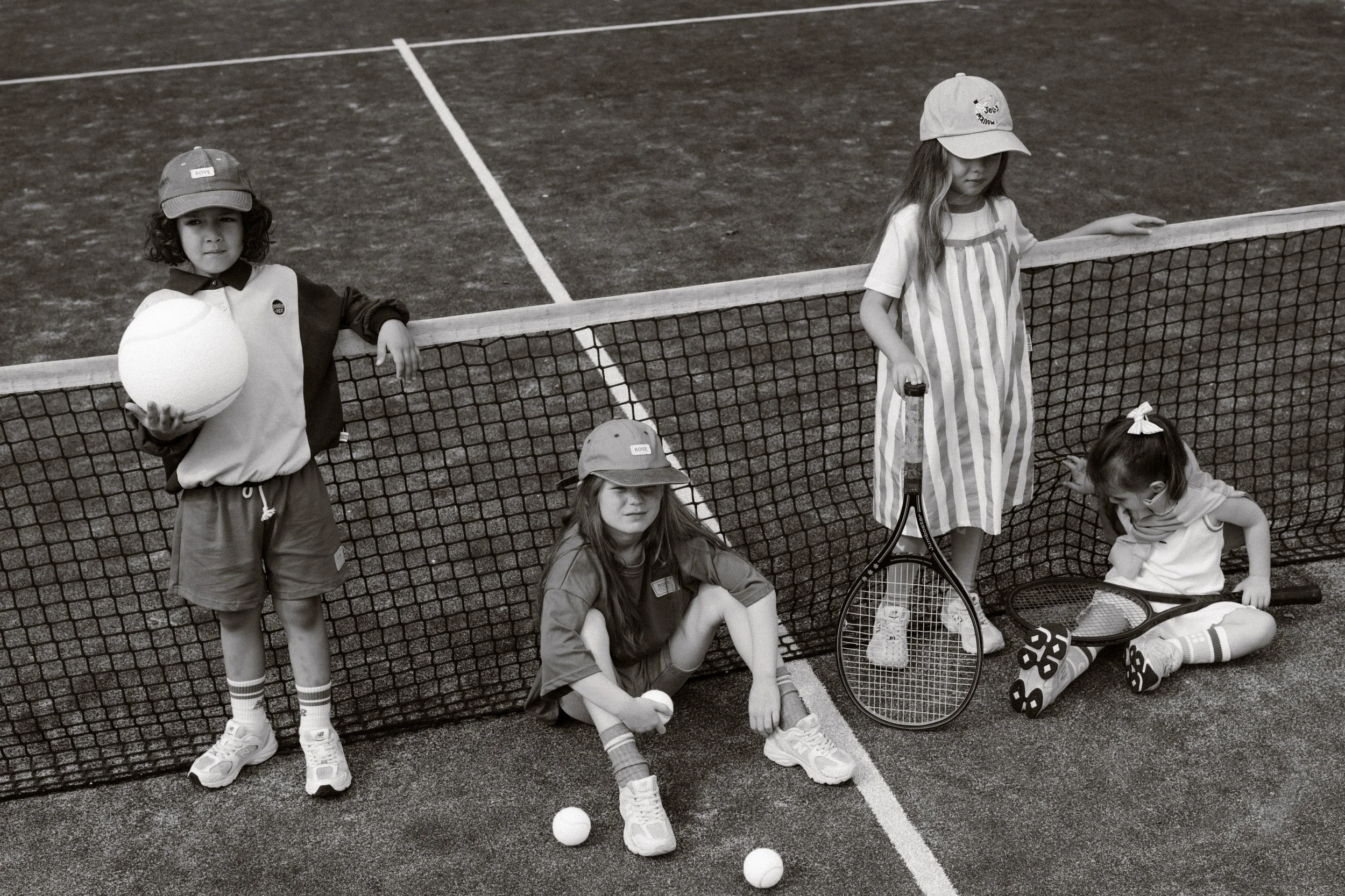 Four children on a tennis court, two standing and two sitting, with tennis rackets, tennis balls, and a tennis net. They are dressed in casual sportswear, some wearing hats. fashion shoot, lifestyle shoot, brandshoot, lieneloe fashion haarlem