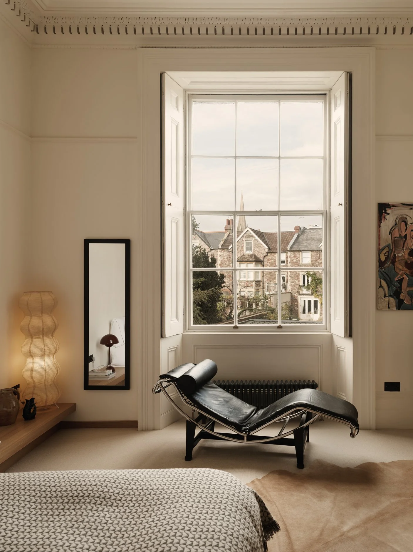 A bedroom with a large window showing houses and a church steeple outside. There is a black leather chaise lounge in front of the window, a tall mirror on the left wall, a bedside lamp, and artwork on the walls.