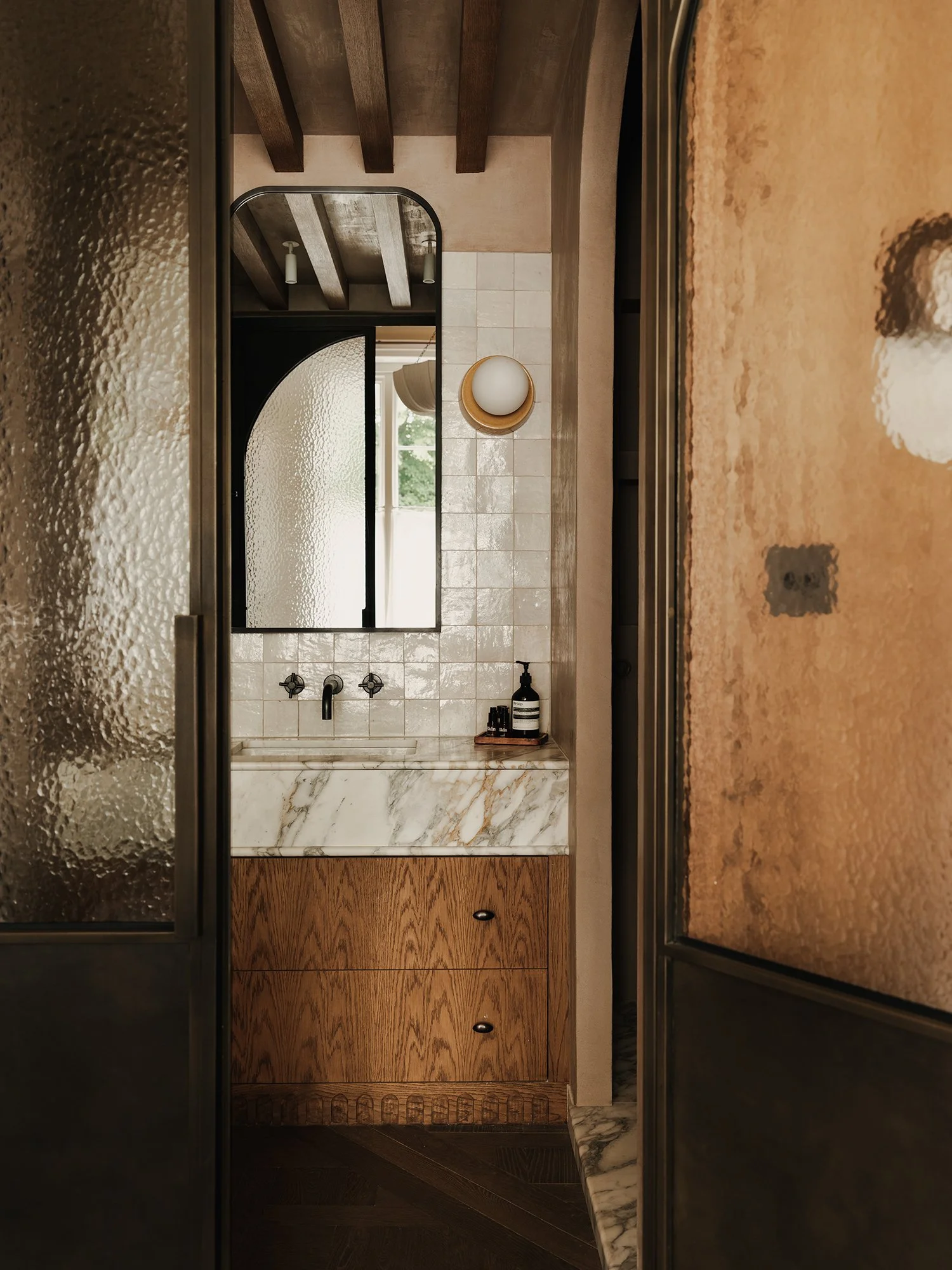 View of a small bathroom sink area with a marble countertop, wooden cabinet, large mirror, and wall-mounted light fixture. Seen through a frosted glass door, with dark wooden ceiling beams and natural light coming from a window.